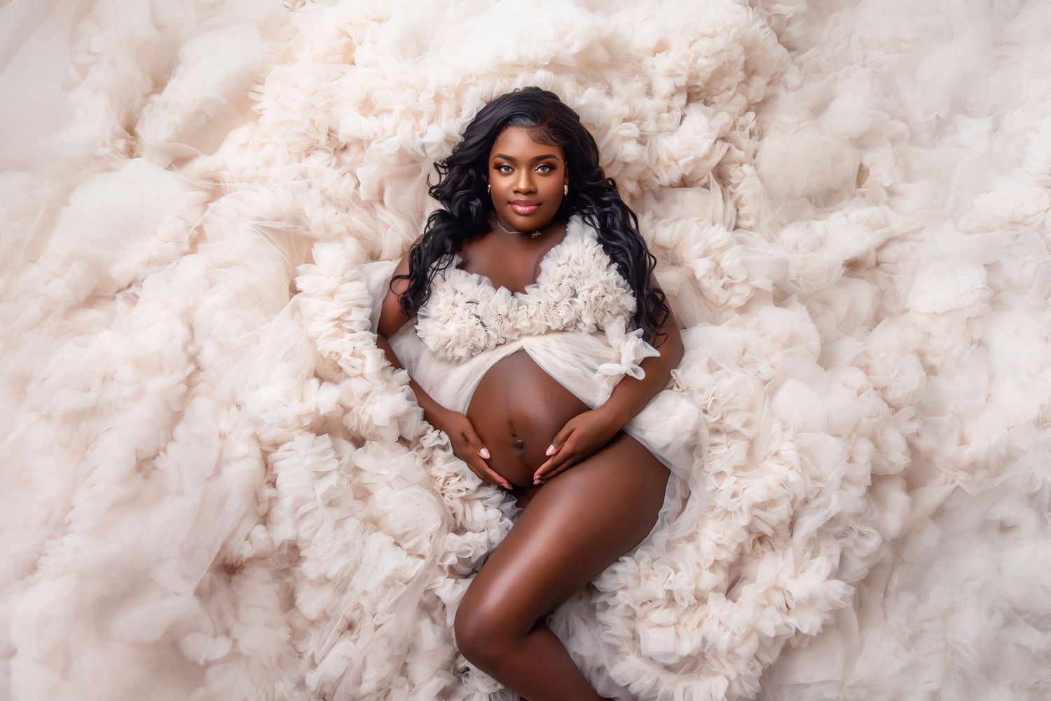 Pregnant woman in fluffy cream wrap poses gracefully on soft beige fabric during a maternity photo shoot.