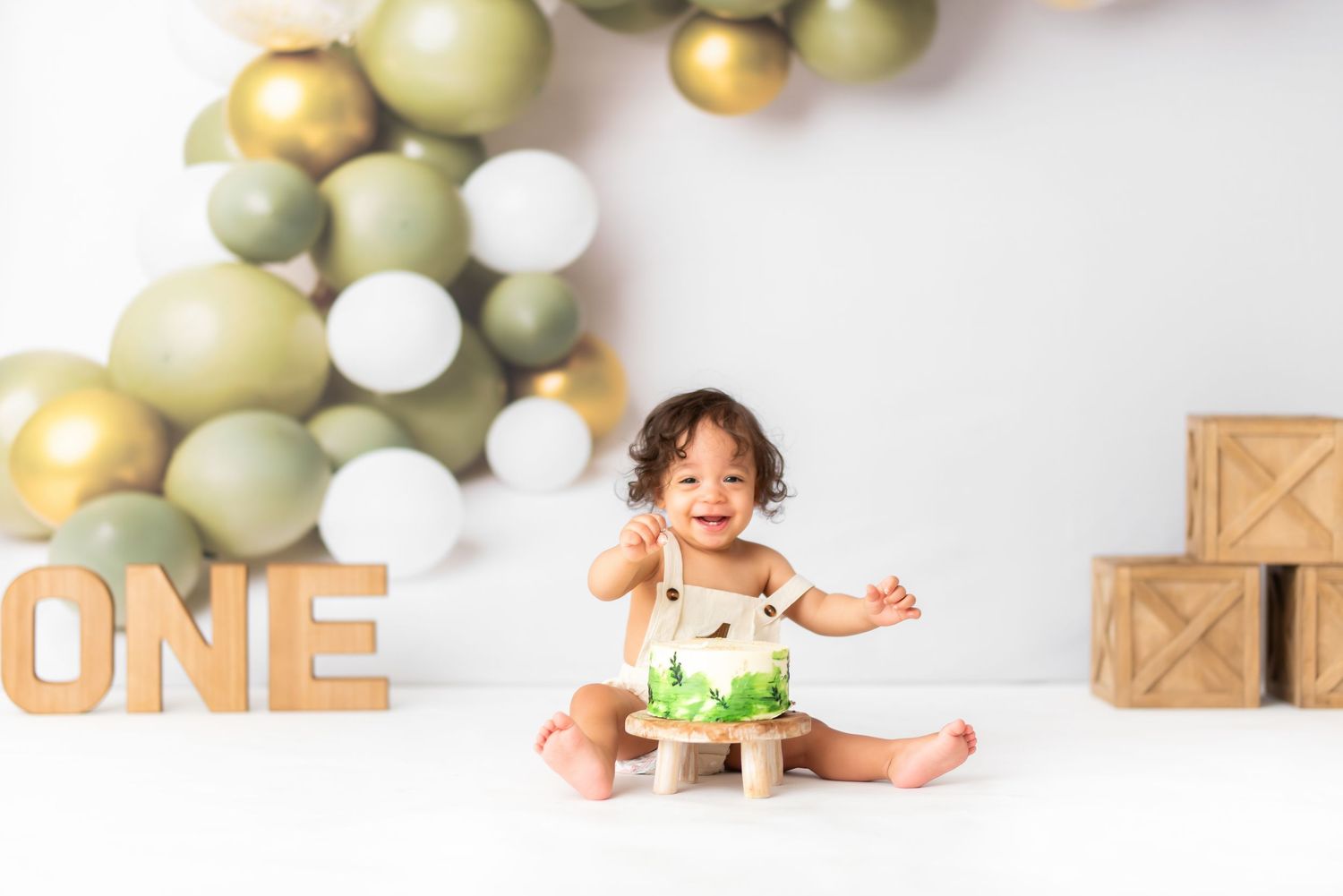 Minimalist cake smash setup with green and gold balloons against white backdrop.