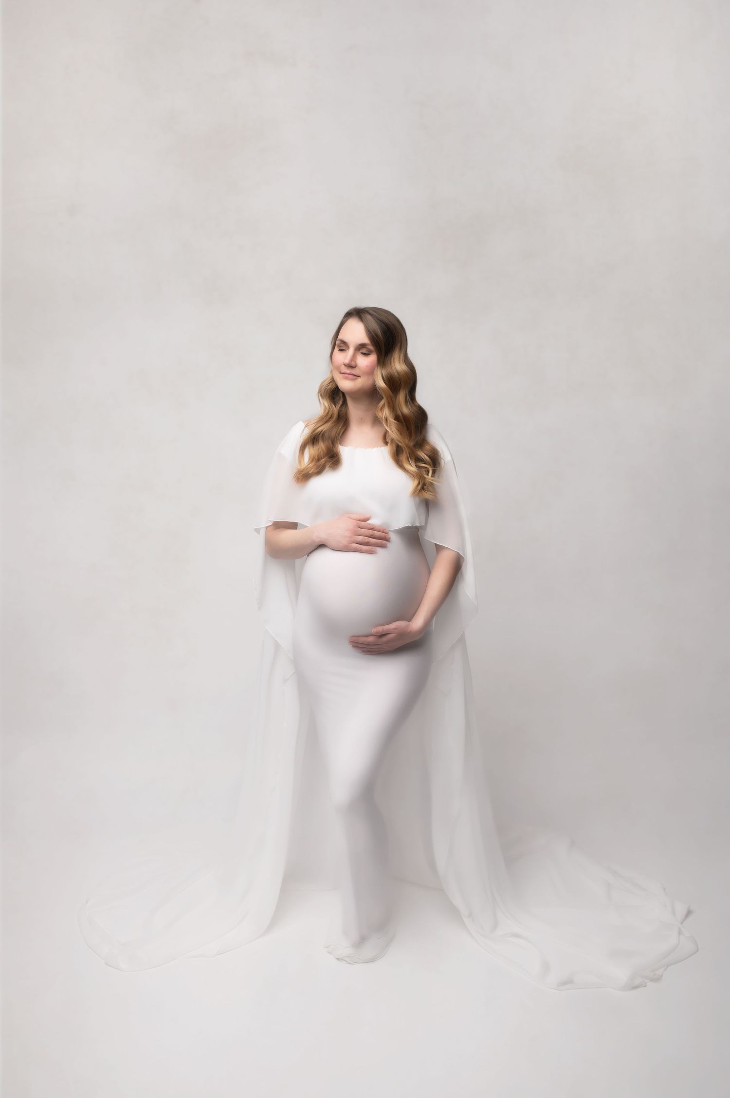 Classic maternity portrait in all white against light grey backdrop with flowing fabric details.