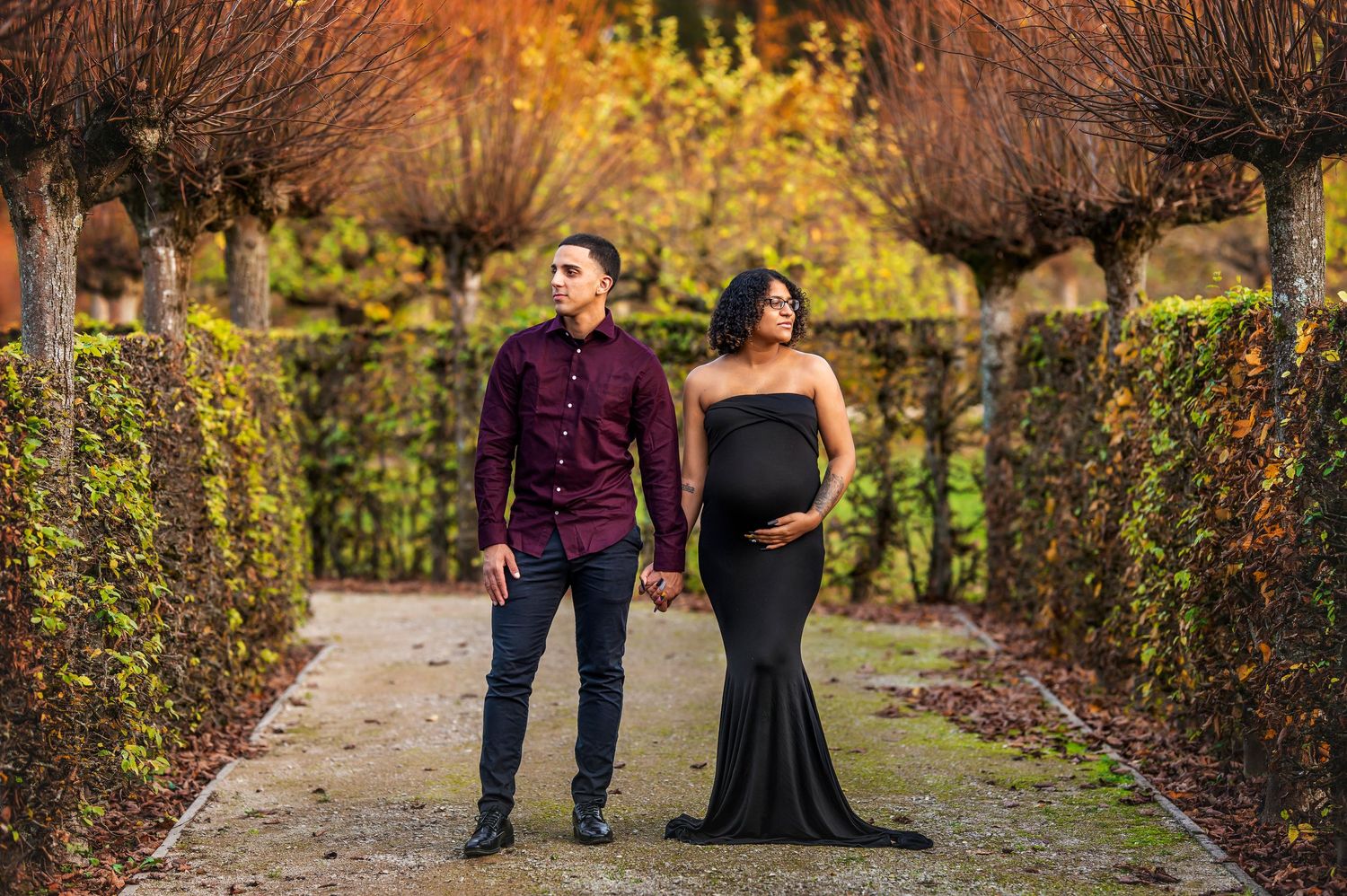Autumn maternity photo of couple walking through garden path with formal attire.
