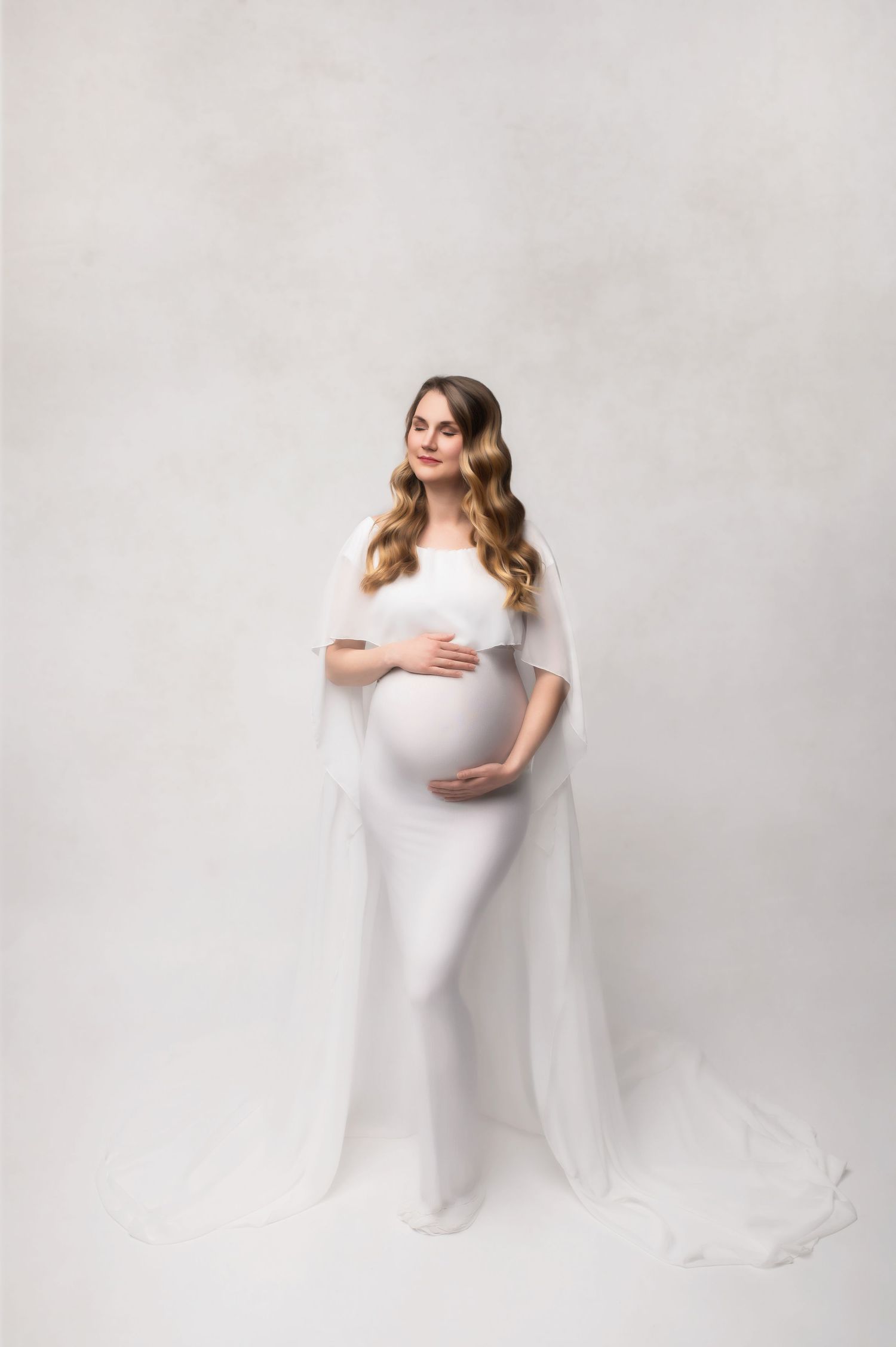 Pregnant woman in elegant white cape gown cradling baby bump during professional maternity photo session.