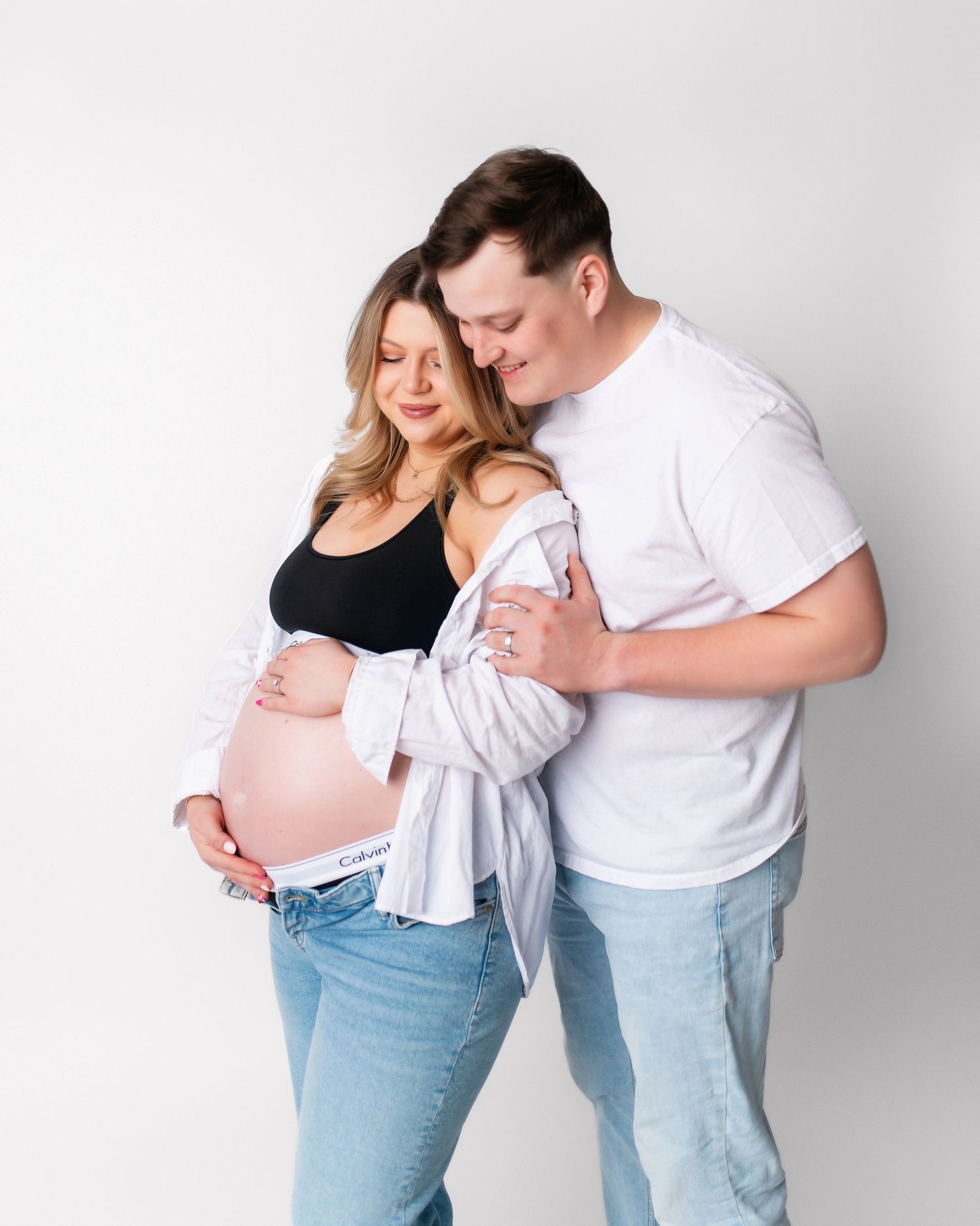 Expectant couple in casual wear share an intimate moment against white background.
