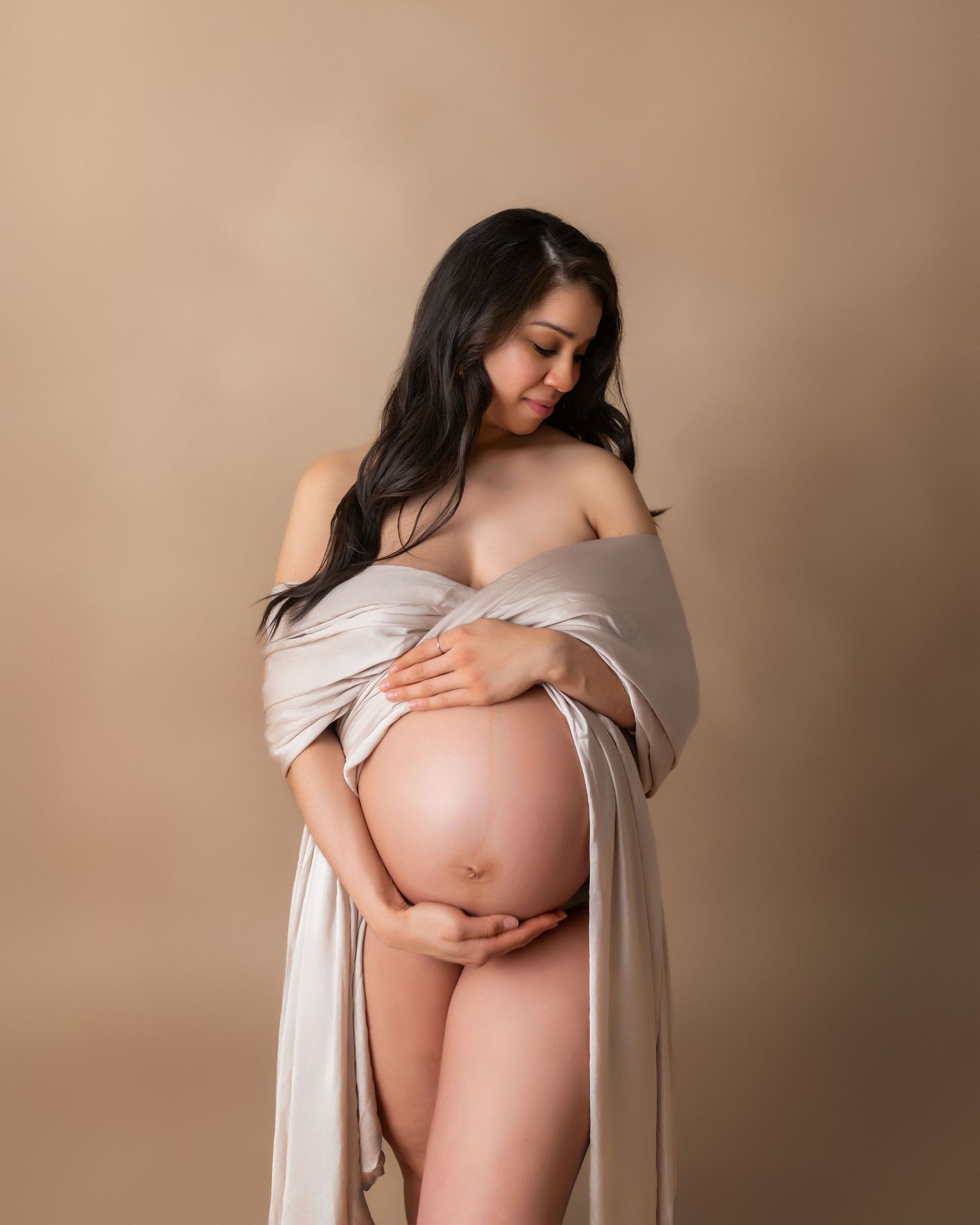 Artistic maternity portrait with woman in draped fabric against neutral background.