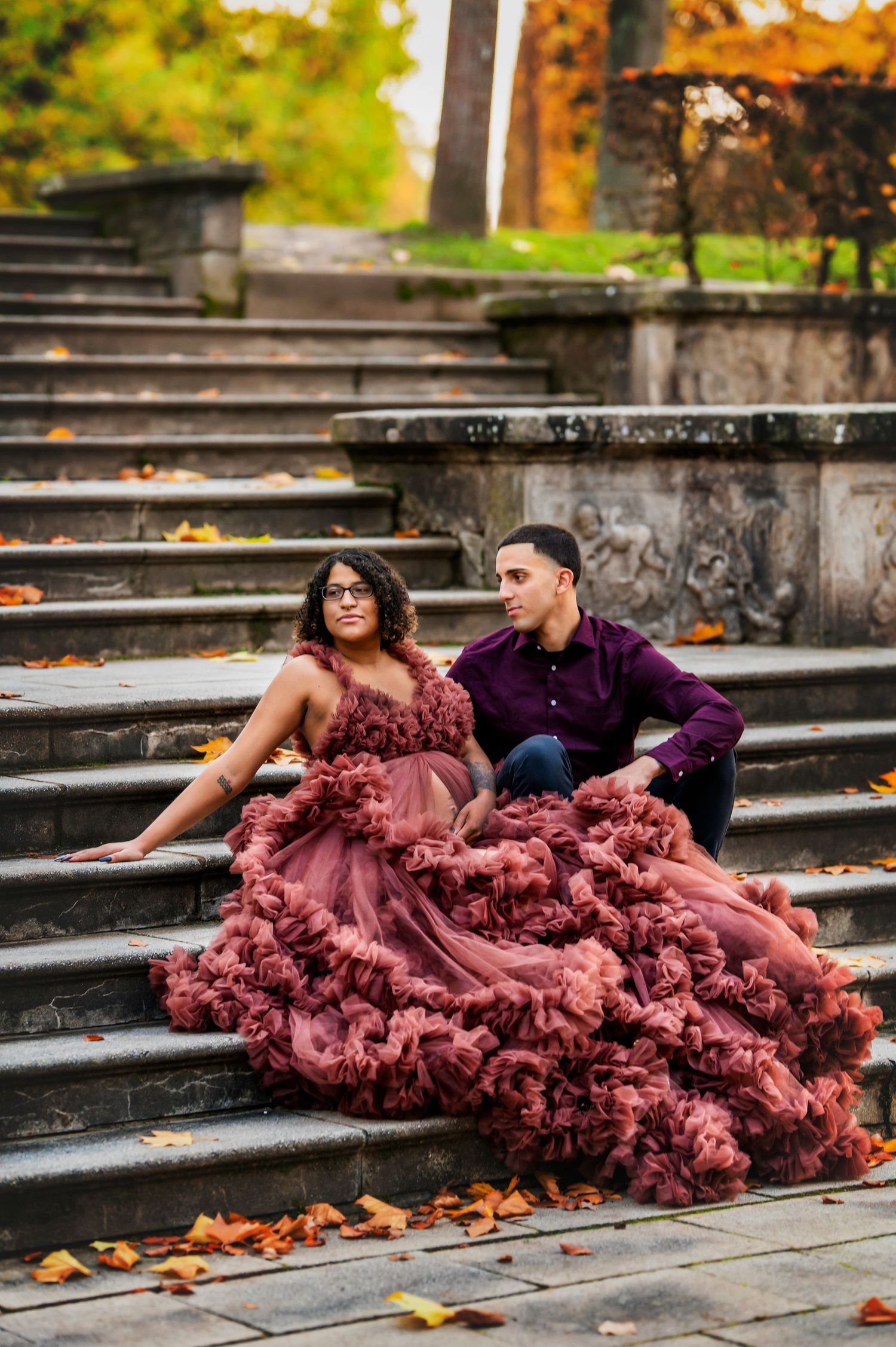 A dramatic fashion photo of a couple on stone steps with a flowing mauve ruffled gown amid autumn leaves.
