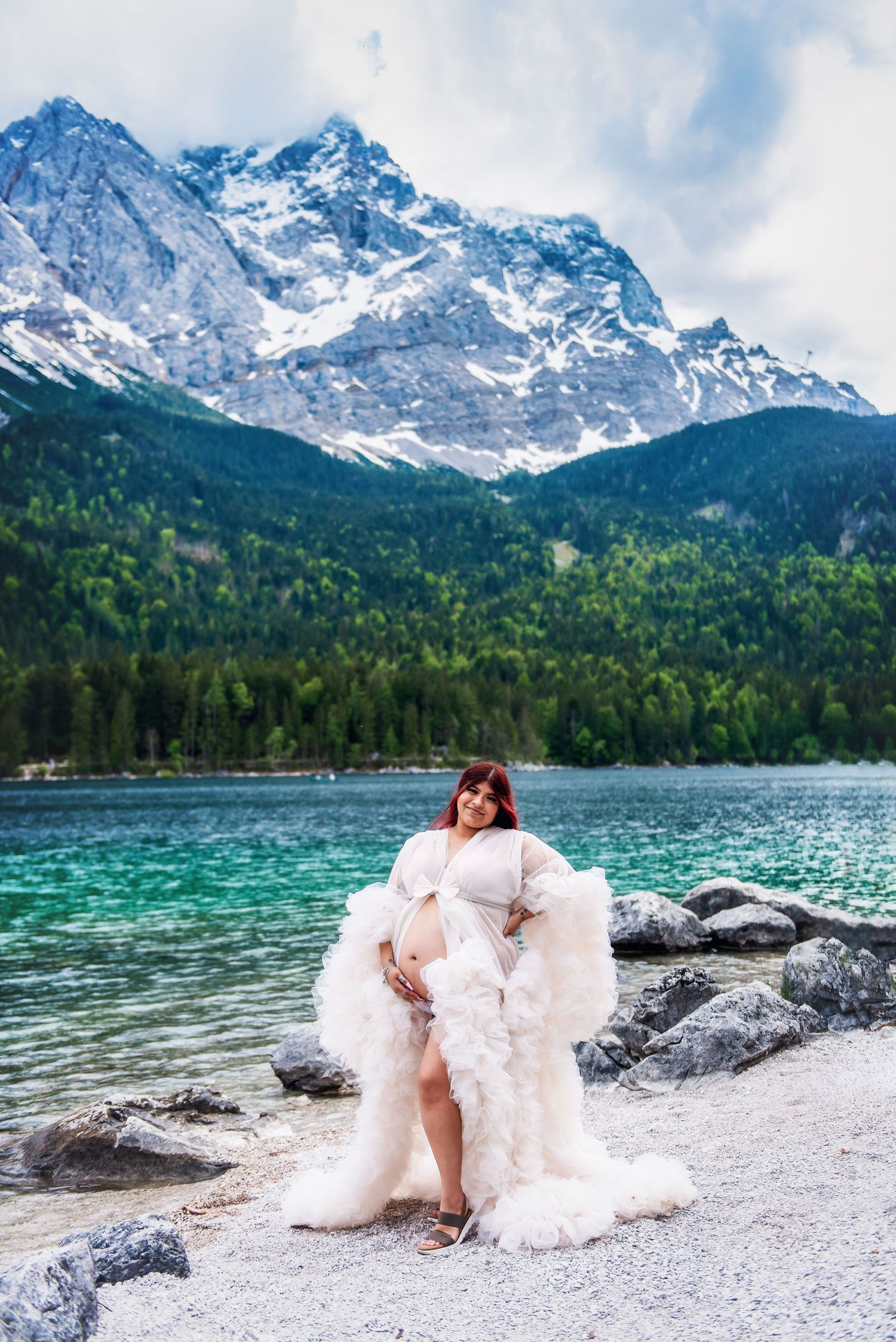A scenic mountain lake landscape with snow-capped peaks and a person in a flowing white dress posing on the rocky shore.