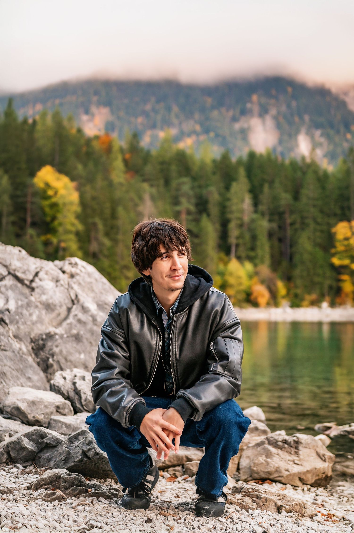 A person in a black jacket sits on rocky lakeside terrain with mountains and autumn forest in the background.