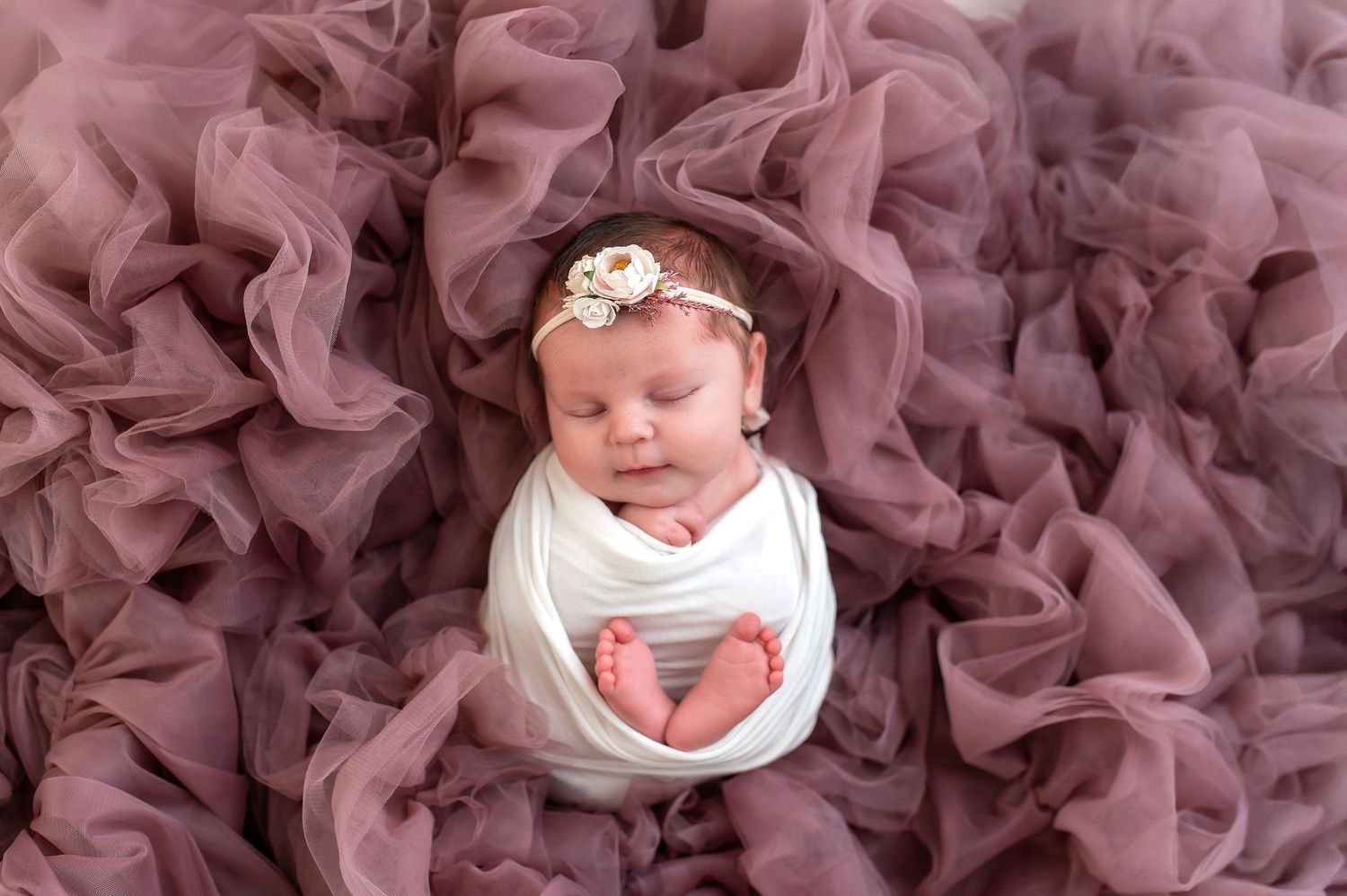 A sleeping baby wearing a white onesie and floral headband lies on a plush mauve tulle backdrop.