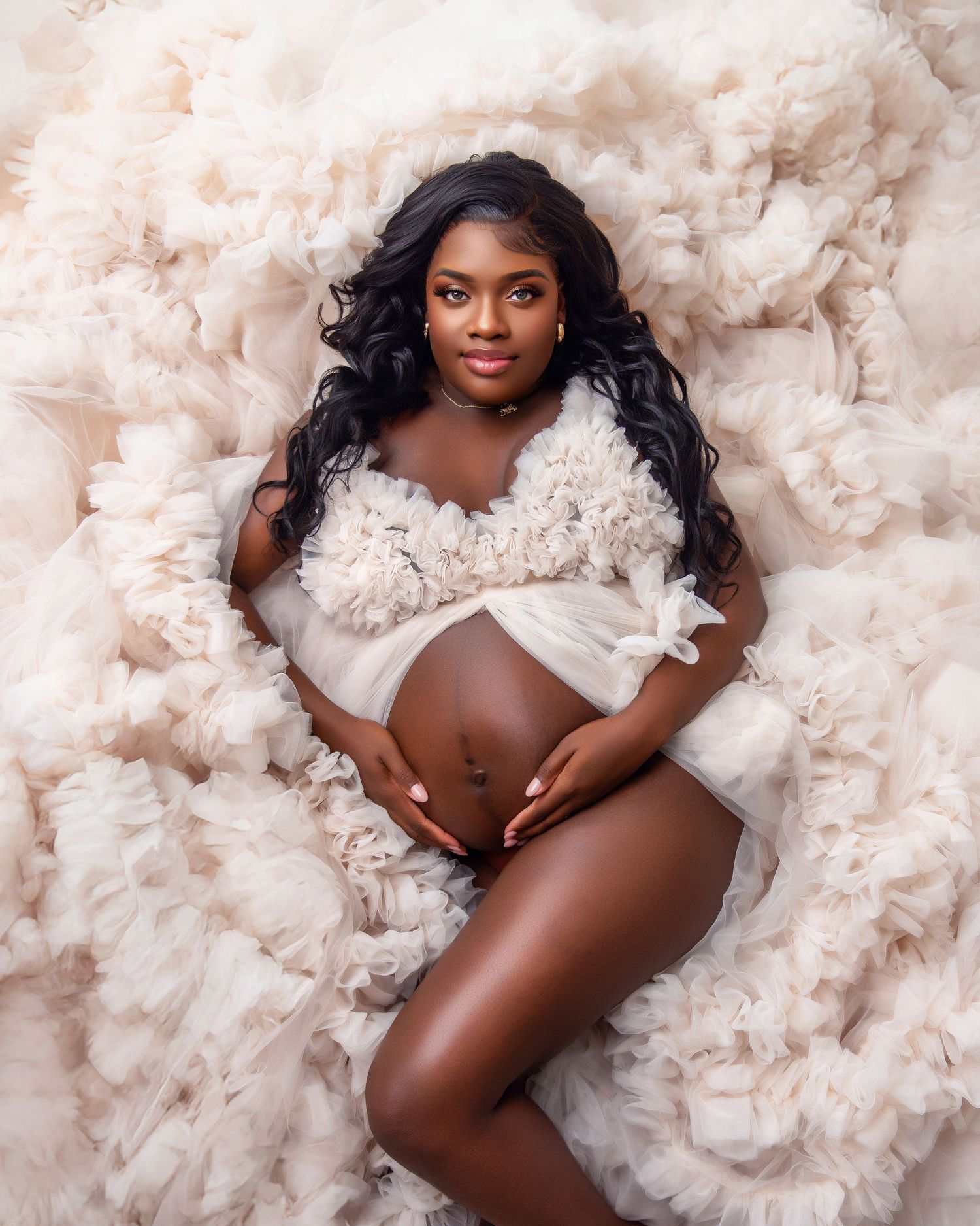 Pregnant woman in ruffled white tulle gown poses gracefully for a maternity photoshoot on a soft floral backdrop.