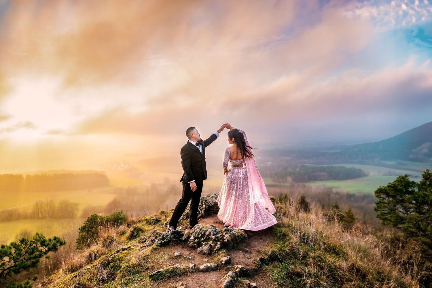 Couple dancing on a scenic hilltop during golden sunset with mountains in the background.