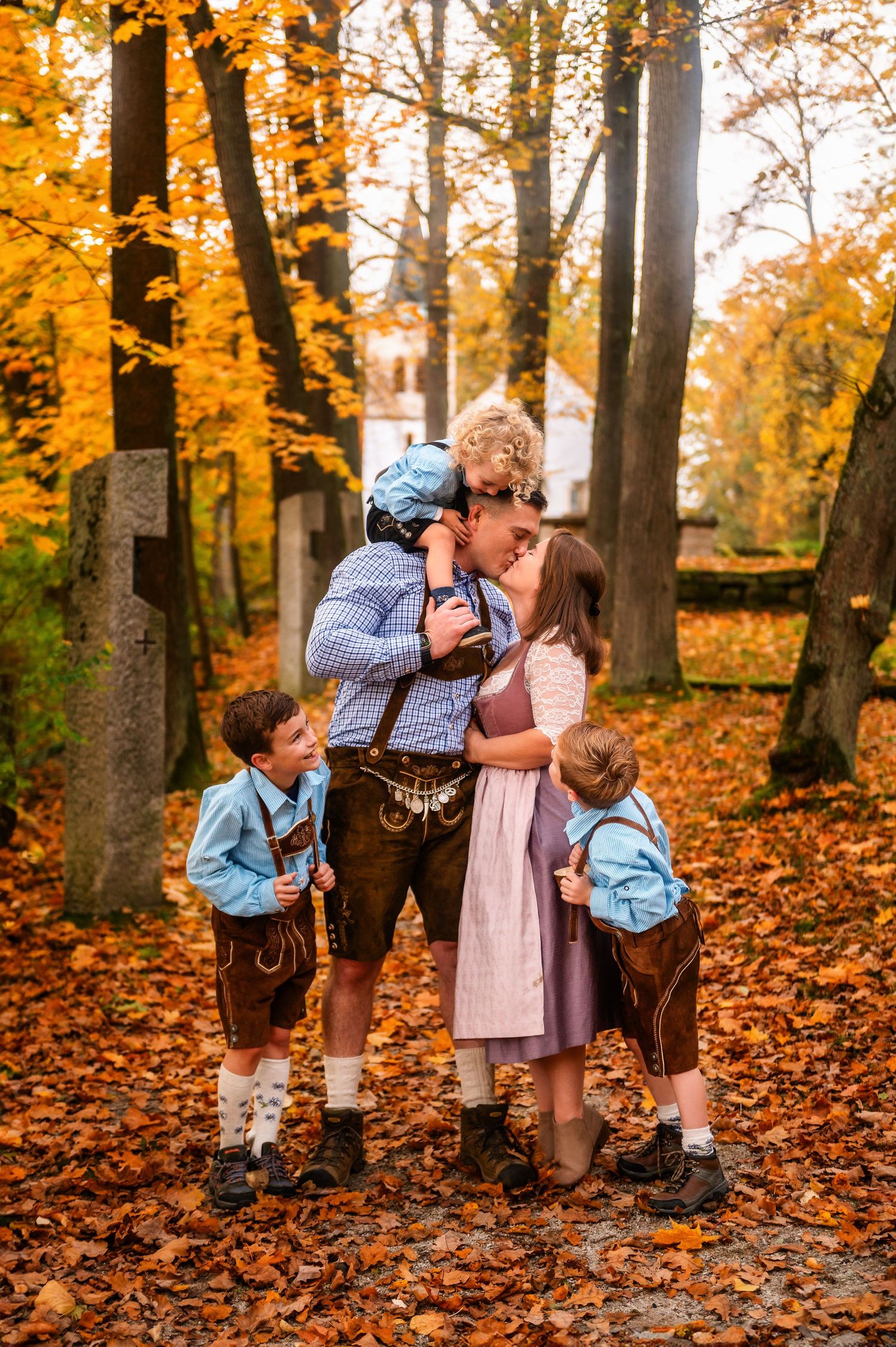 Couple embraces on path covered in yellow autumn leaves while child in traditional dress looks on.