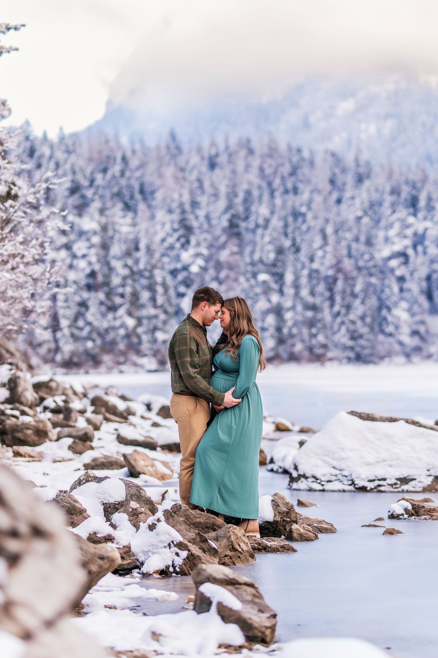 A couple shares an intimate moment on snow-covered rocks with a snowy forest and mountains in the background.