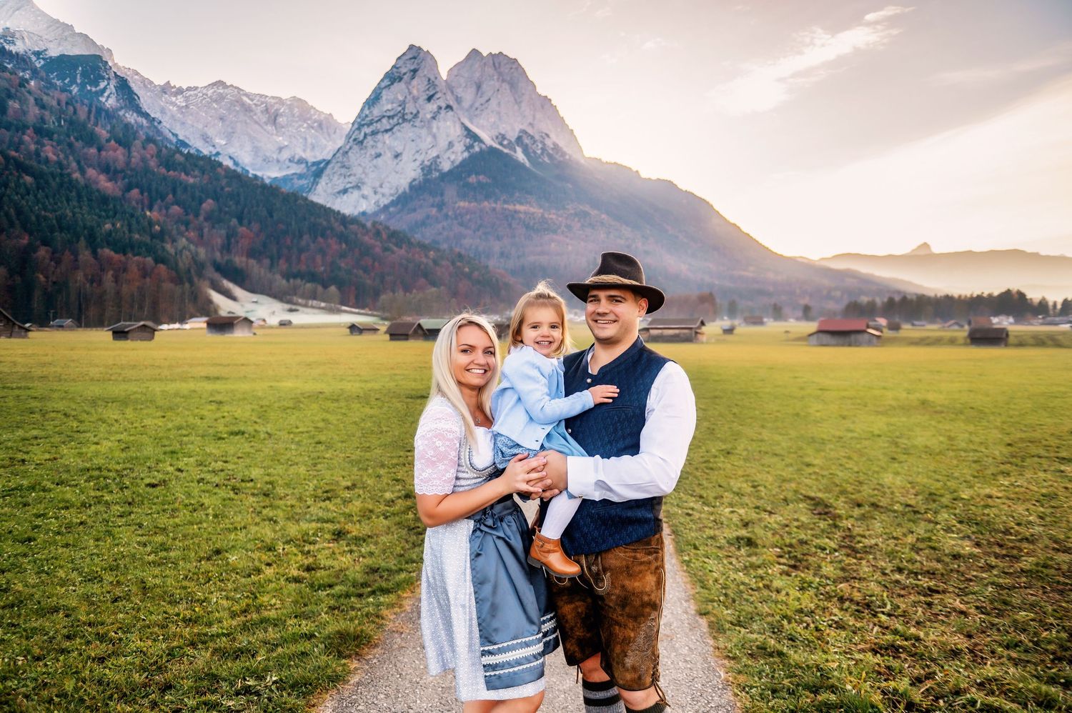 Traditional Bavarian family poses in front of majestic Alpine mountain peaks in Garmisch-Partenkirchen meadow at sunset.