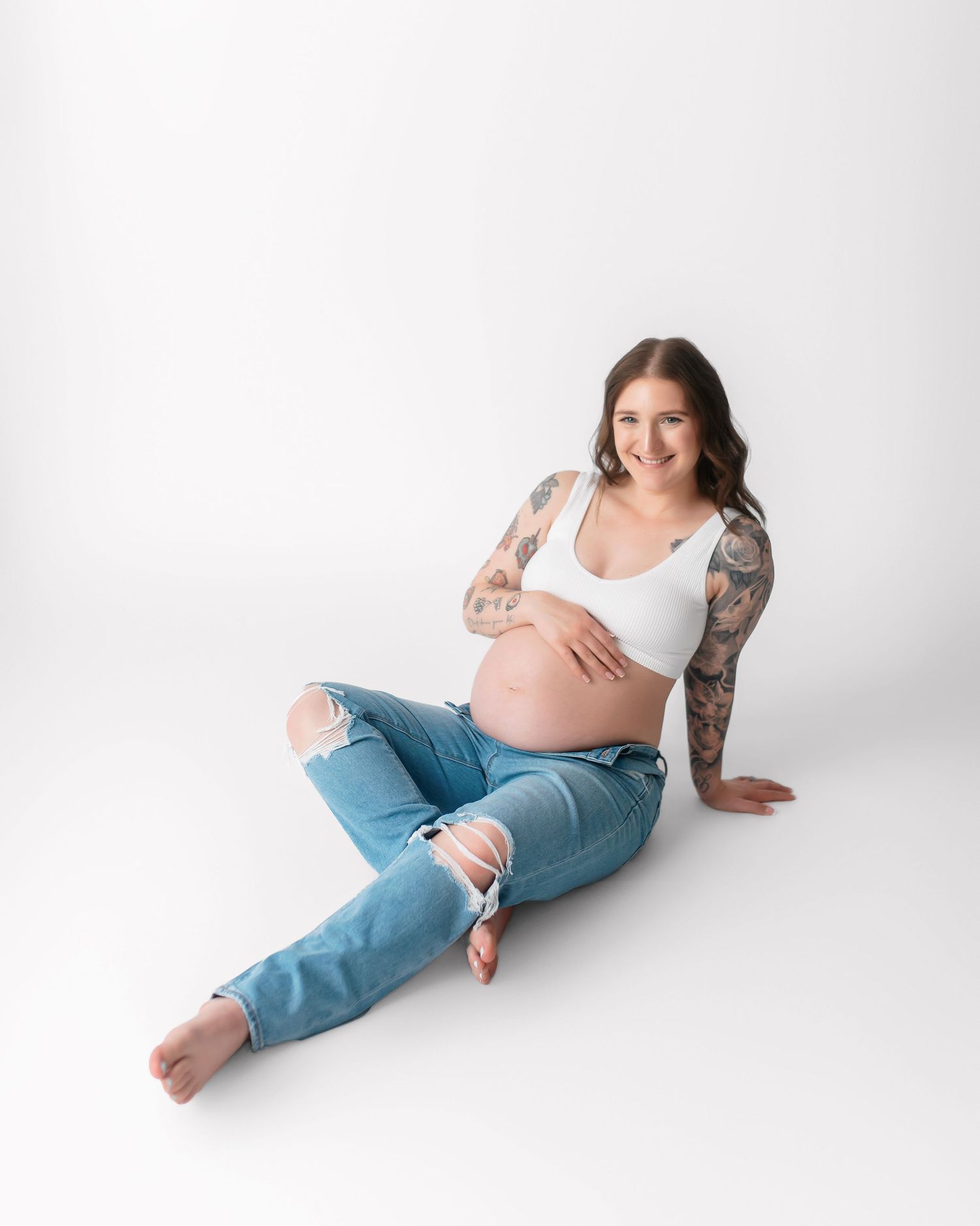 Expectant mother with tattoos poses casually in distressed jeans against white backdrop.