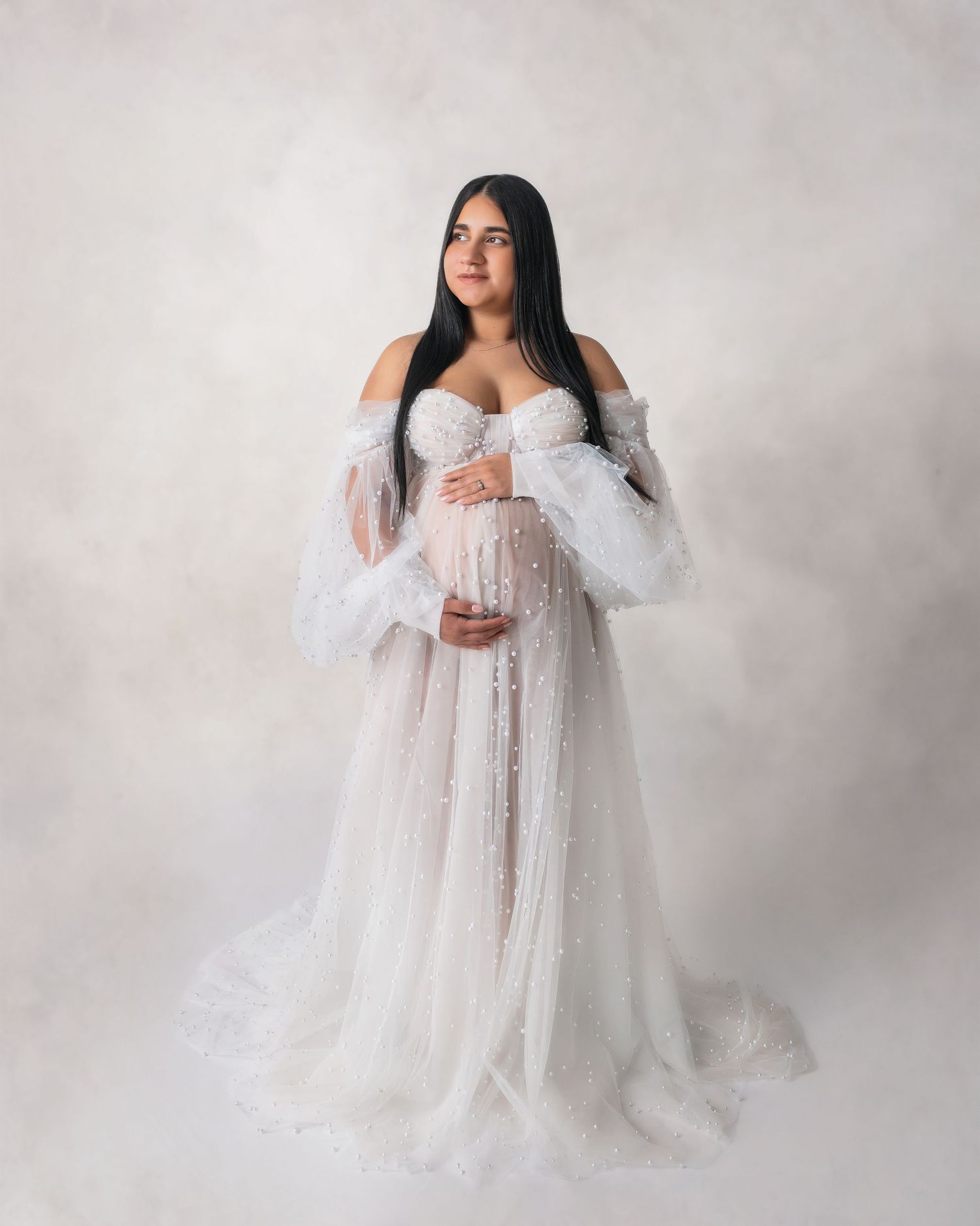Pregnant woman in elegant white off-shoulder tulle maternity gown cradling baby bump in professional studio photo.