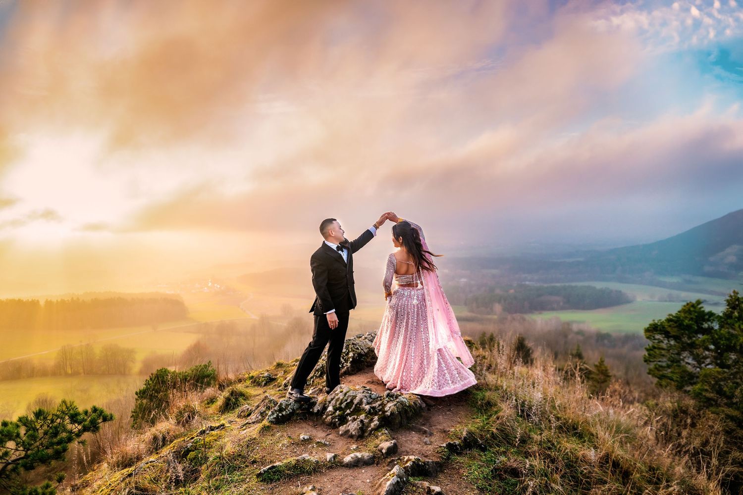 Couple dancing on mountain top at sunset, woman in pink gown, man in black suit, scenic valley backdrop.