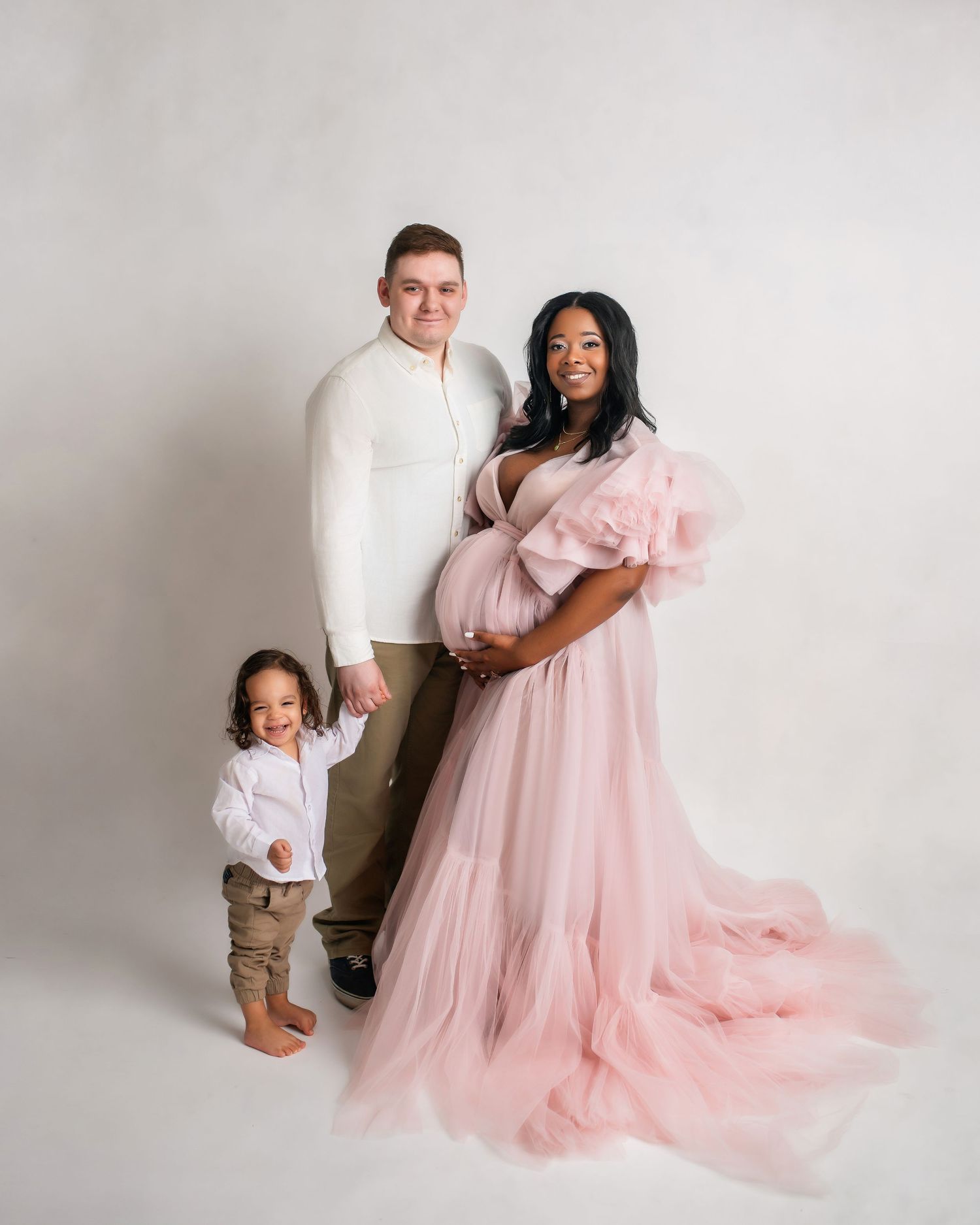 Expecting family of three poses in studio, woman in pink tulle maternity gown, man in white, toddler son beside them.