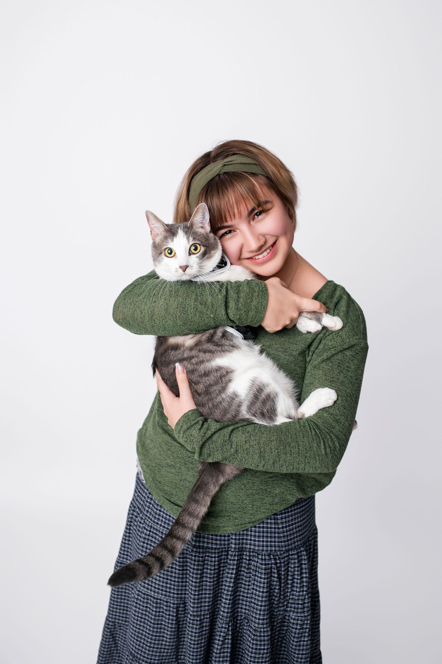 Smiling young woman in green sweater holding a gray and white tabby cat against a white background.