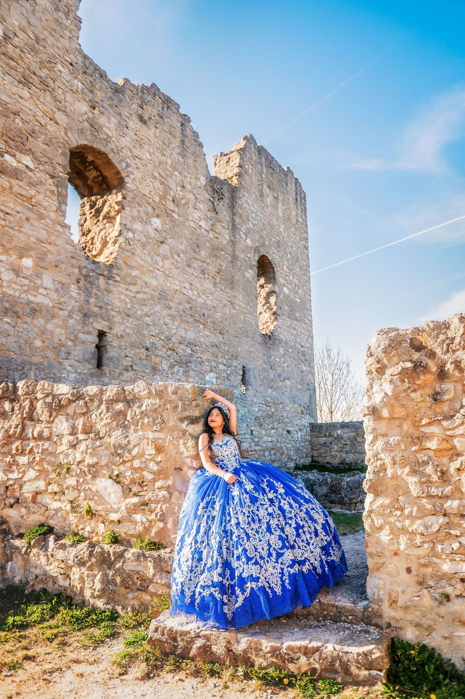 Young woman in blue ball gown poses at ancient stone castle ruins under blue sky.