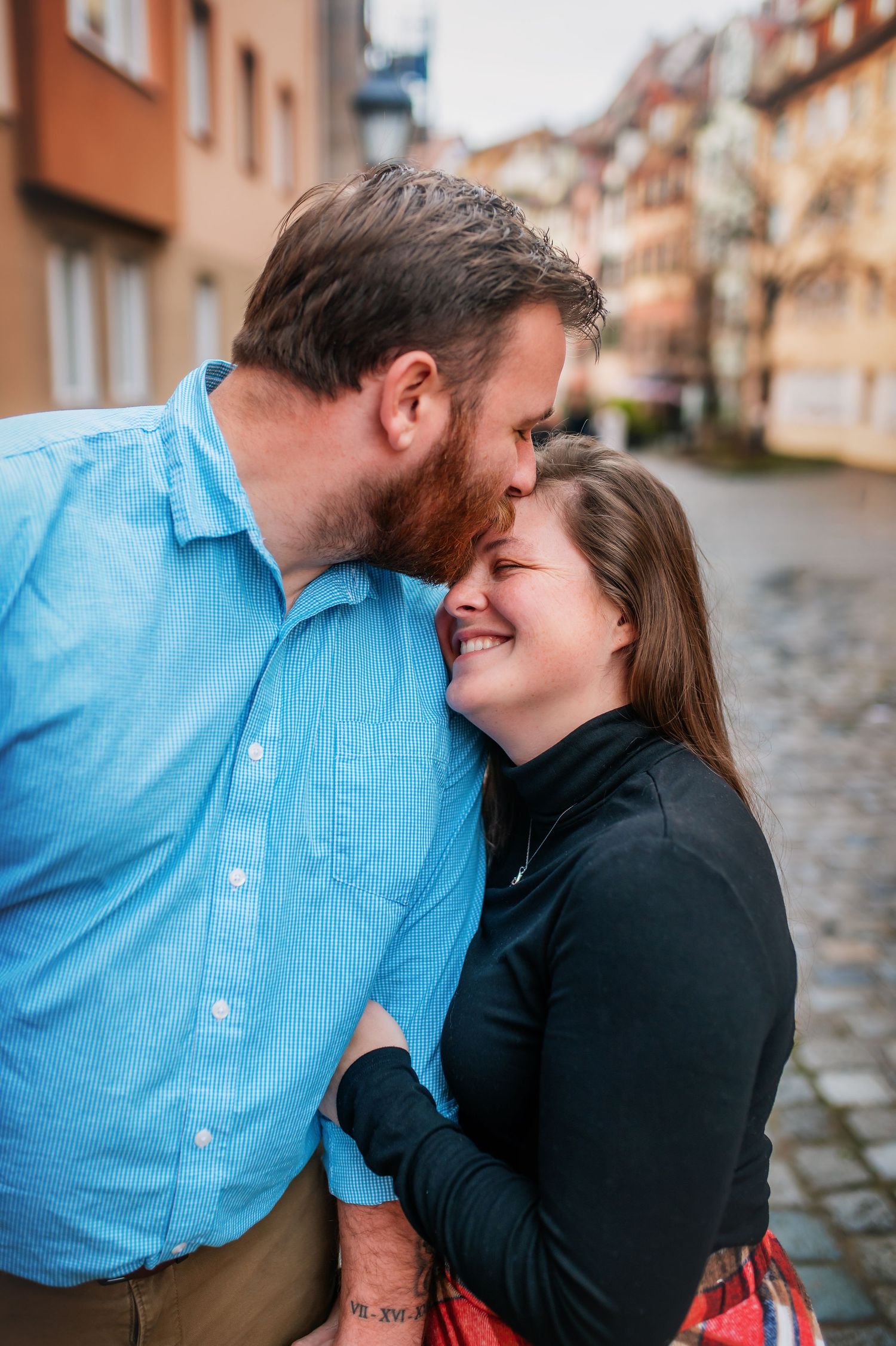 A couple shares an intimate moment while laughing together on a cobblestone street in front of colorful apartment buildings.