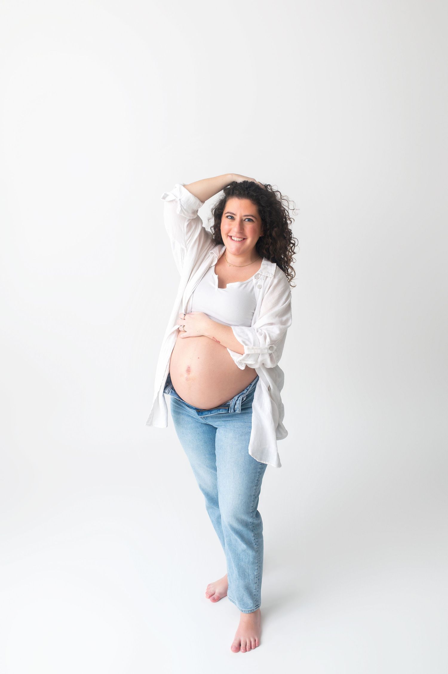 Expecting mother in casual attire poses playfully against white background during maternity session.