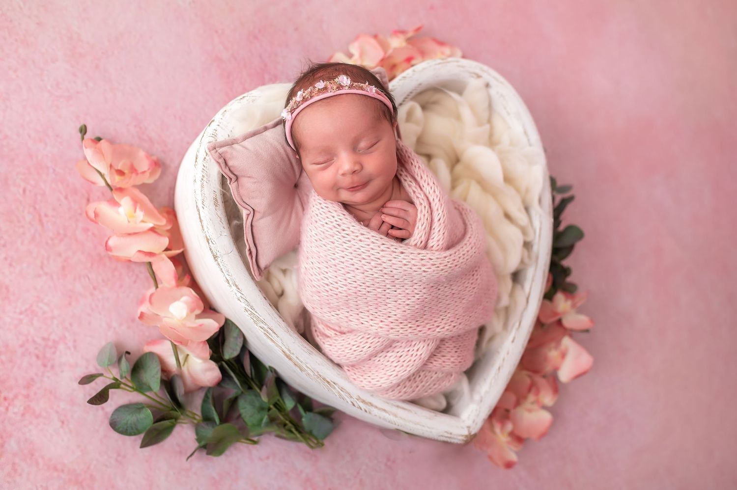 Sleeping newborn posed in heart-shaped basket with pink roses and delicate crown.