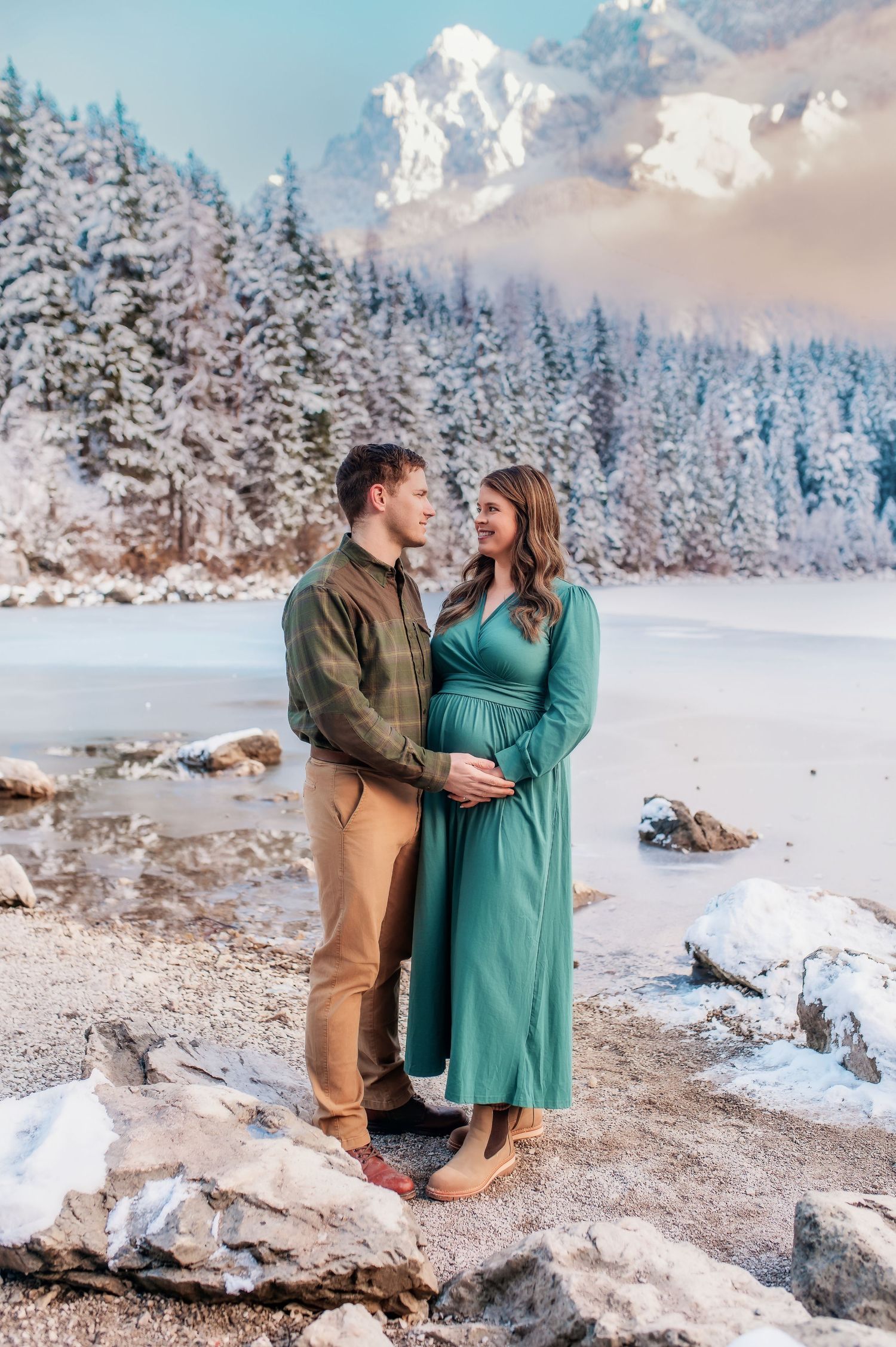 A romantic winter photo shoot at a snow-covered mountain lake with snow-capped peaks and evergreen trees in the background.