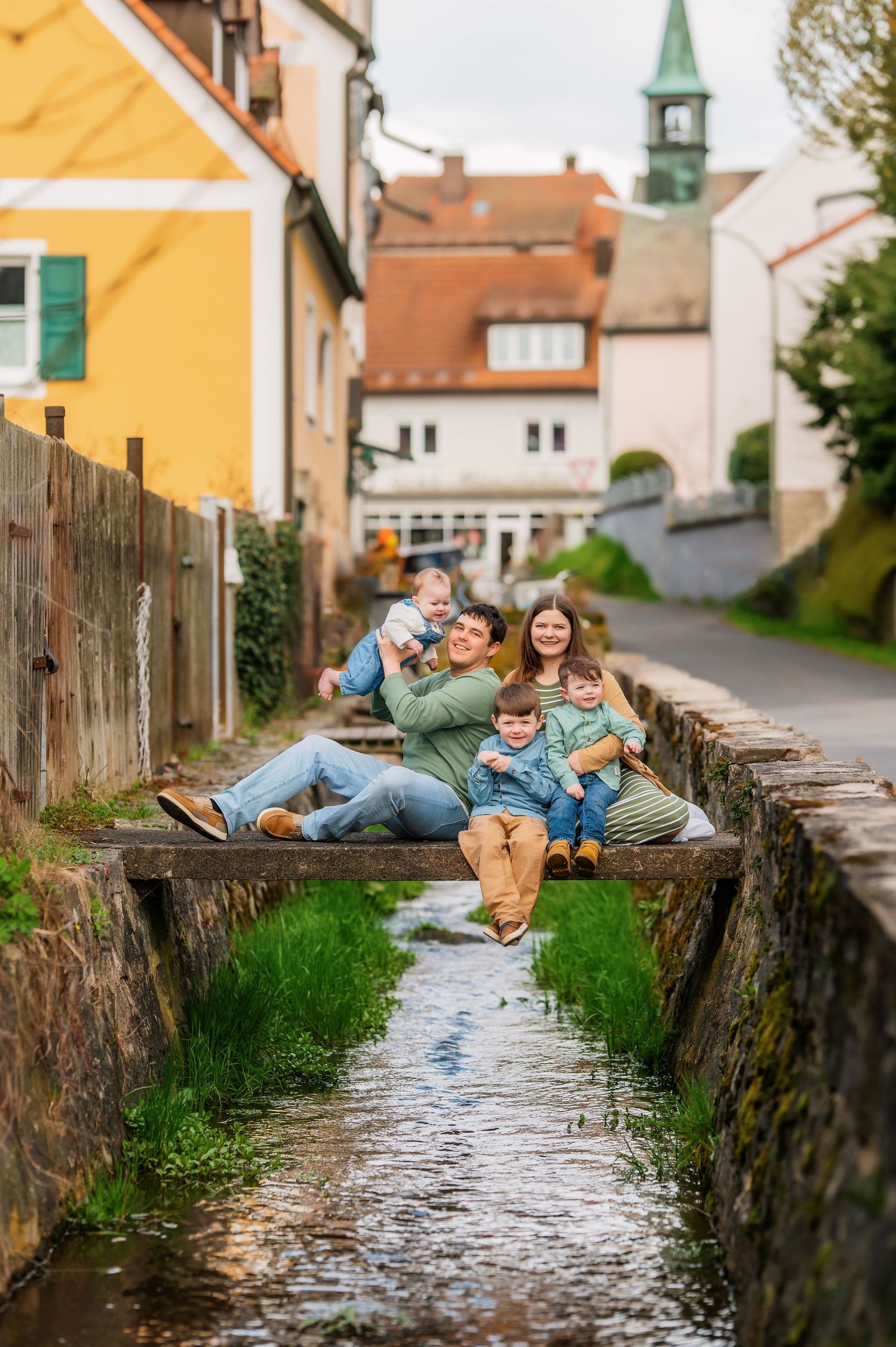 A couple playfully poses along a stone wall with water in a picturesque European village setting.