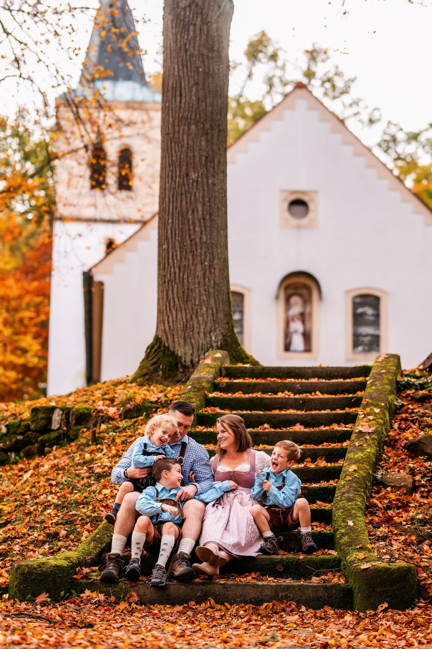 Family rests on moss-covered stone steps near a white church surrounded by autumn leaves.