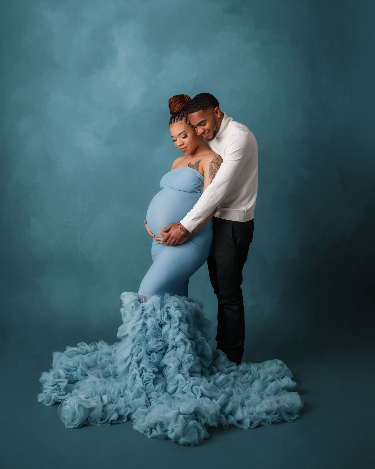 Intimate maternity portrait featuring a couple in blue and white against a teal backdrop with flowing fabric.
