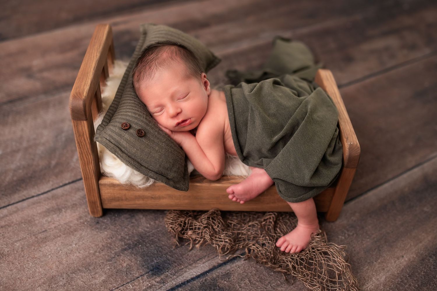 Sleeping newborn posed in wooden bed with green blanket and military accessories.