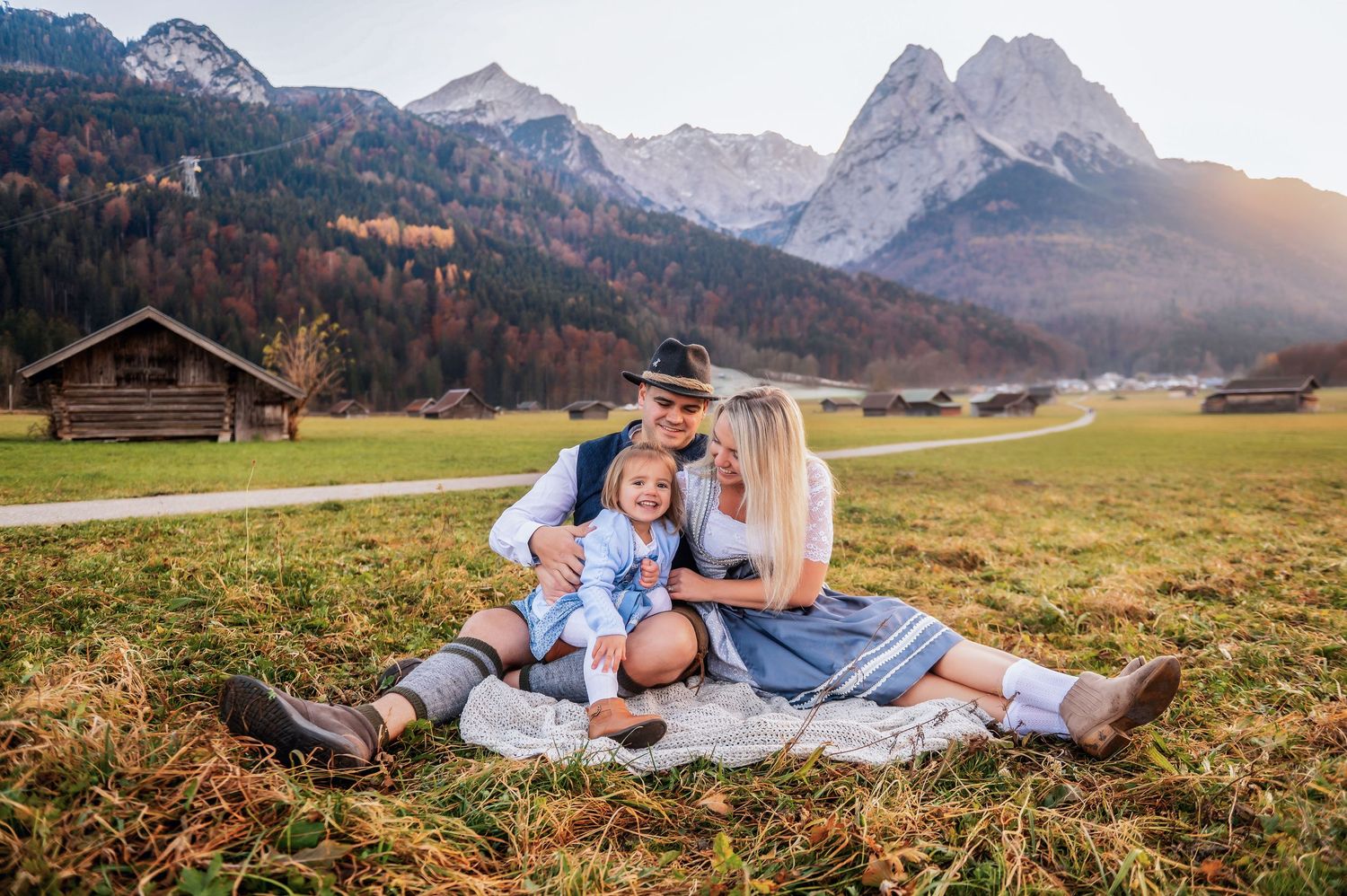 Family in traditional Bavarian attire sitting on a blanket in an alpine meadow with mountain peaks and wooden chalets.