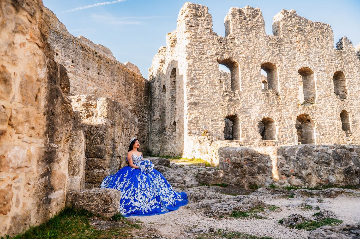 Young woman in a royal blue quinceañera gown poses gracefully among ancient stone castle ruins under a clear sky.