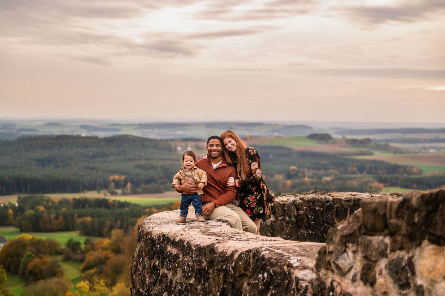 A family sits together on castle ruins overlooking scenic countryside at sunset.