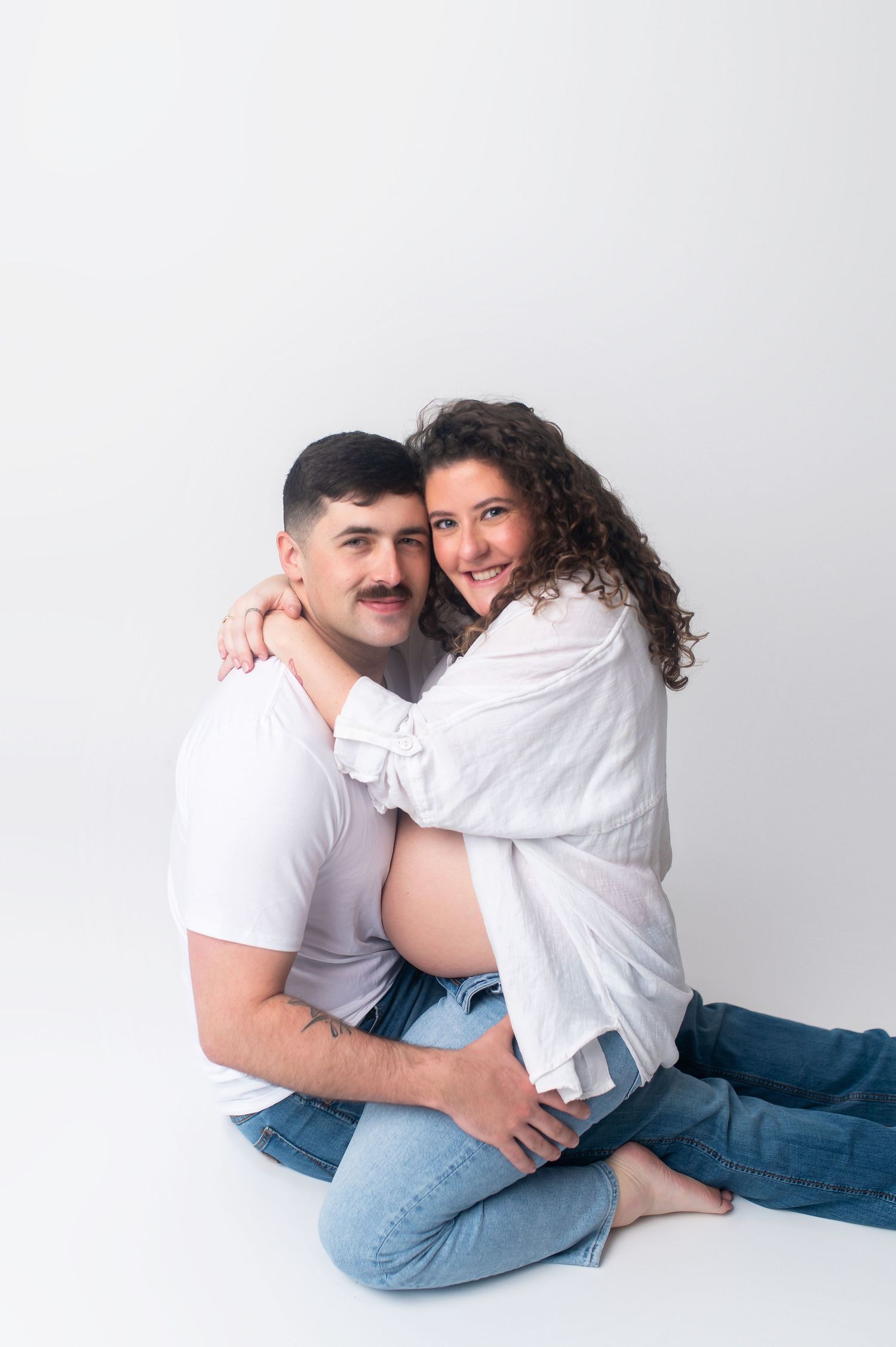 Couple in white tops and blue jeans embrace lovingly against white background during maternity photoshoot.