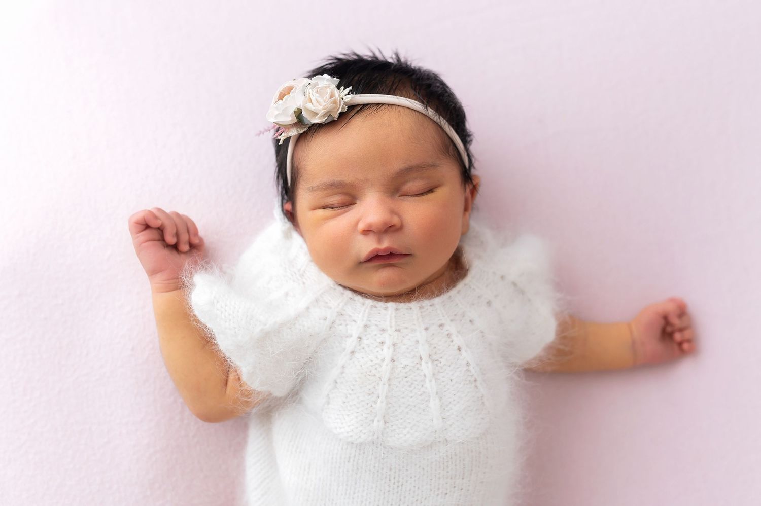 Sleeping newborn wearing white lace dress and floral headband on pink background.
