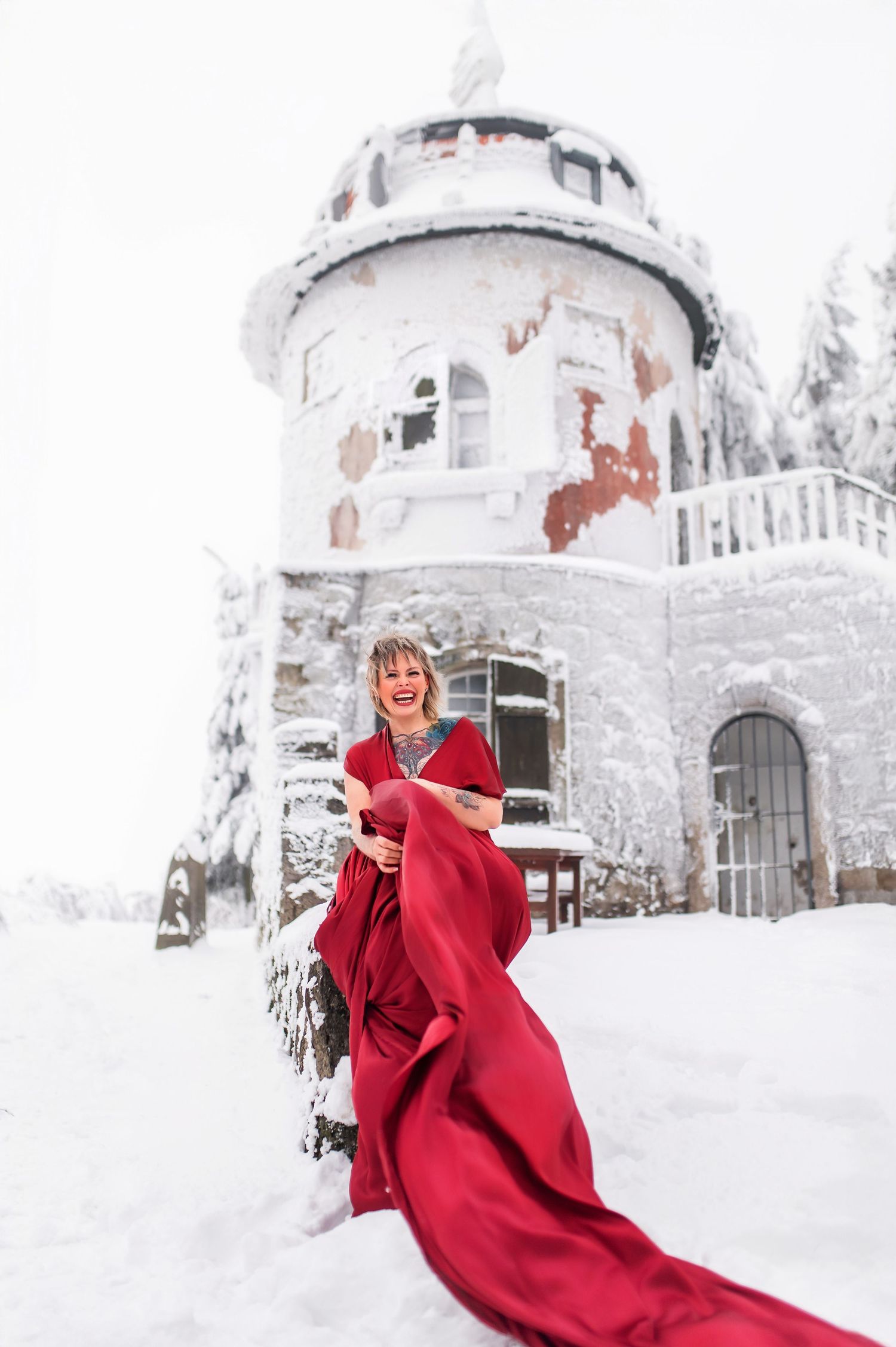 Woman in red gown laughing outside snow-covered castle in winter wonderland setting.