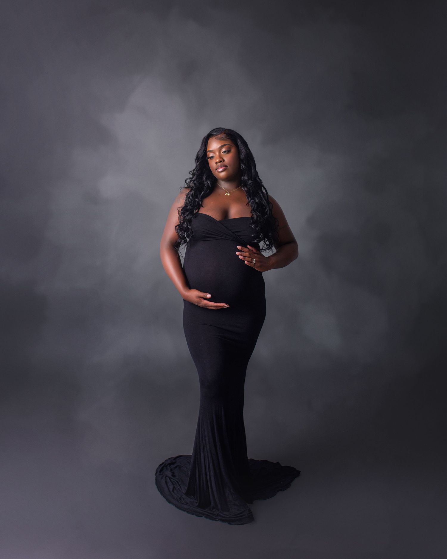 Elegant maternity photo of a woman in a black fitted gown against moody gray backdrop.