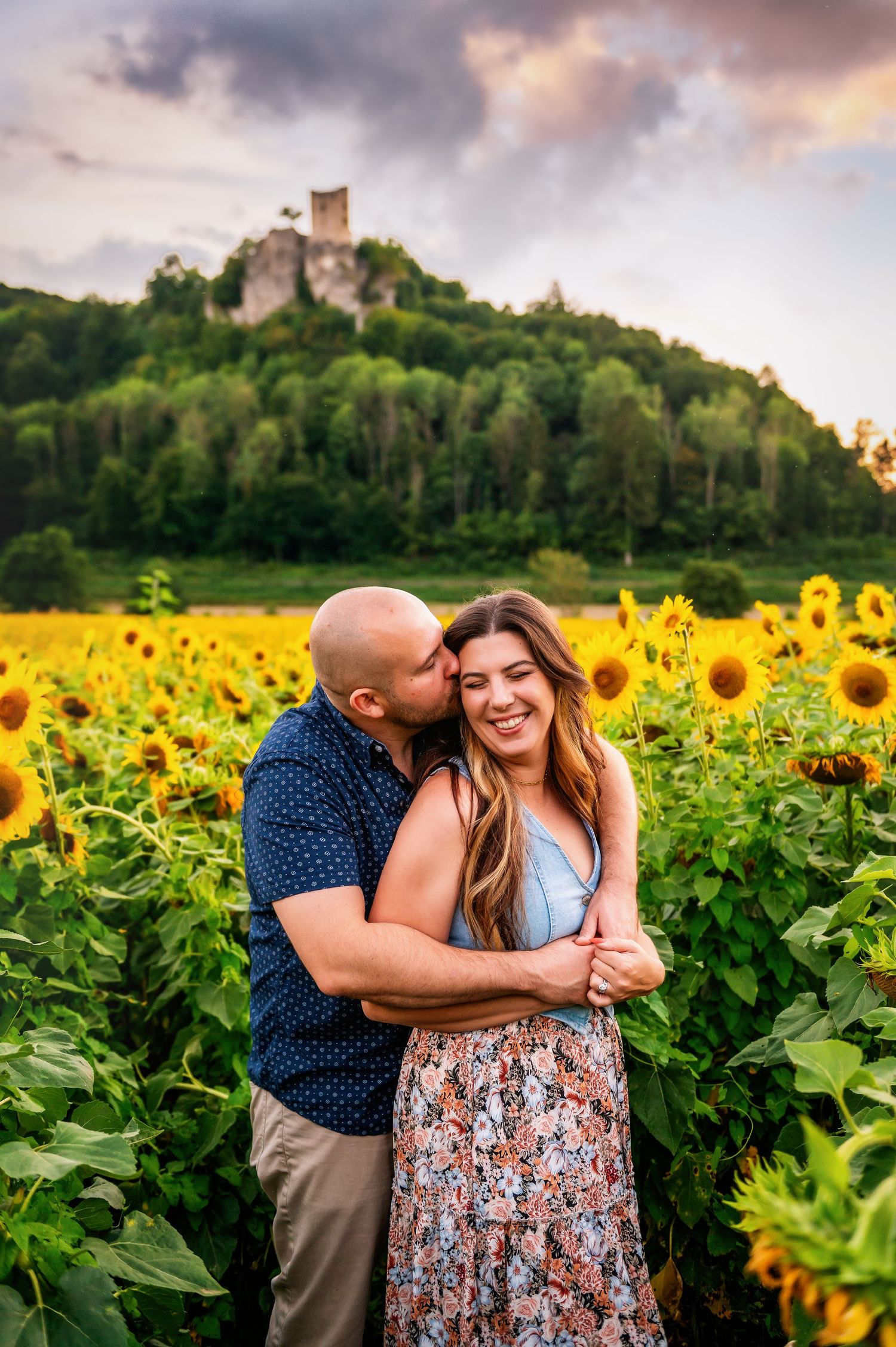A couple shares an intimate moment in a vibrant sunflower field with a castle on a hill in the background at sunset.