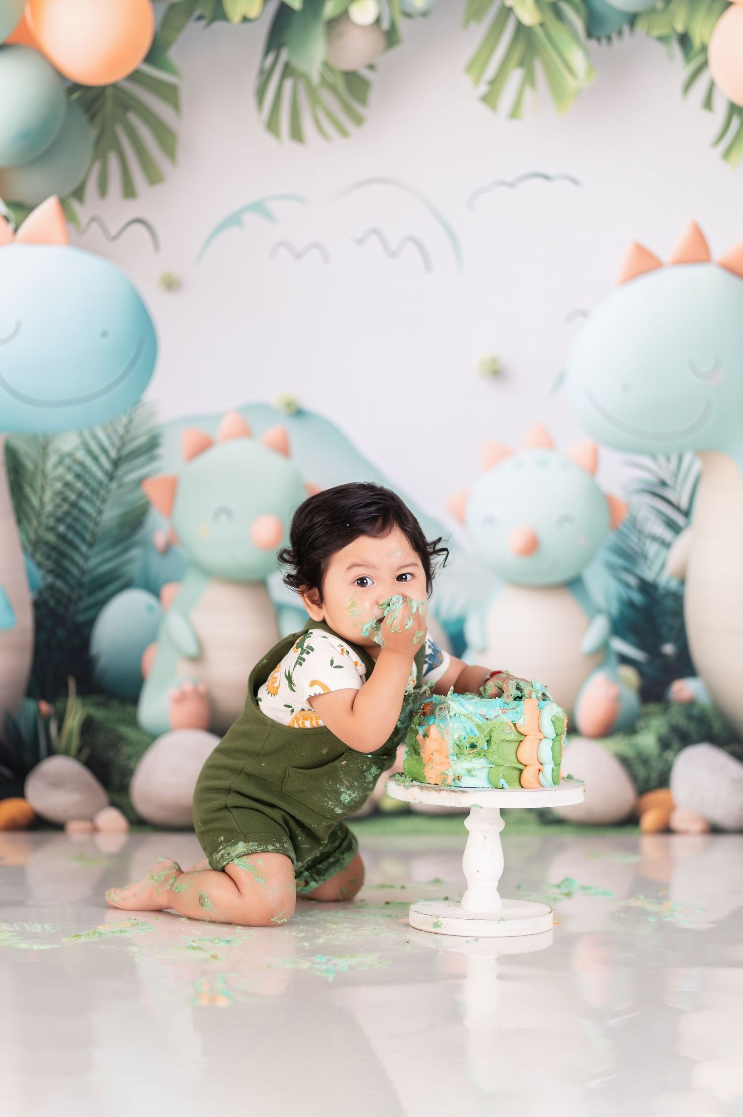 Baby enjoying cake smash photography session with dinosaur themed decorations and balloons.