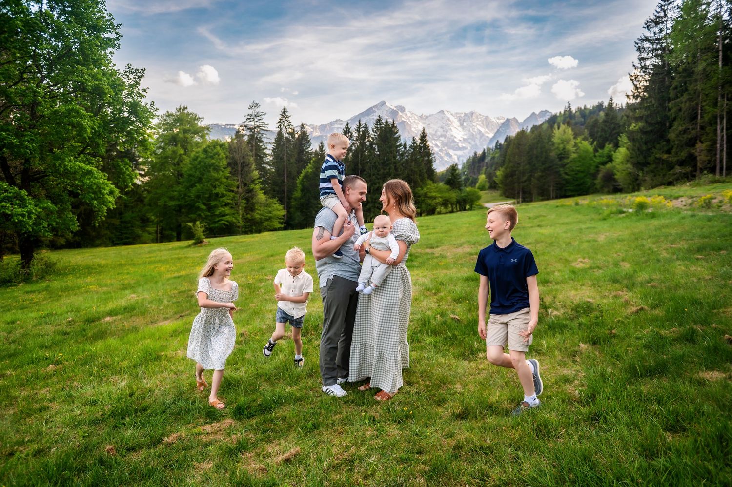 Family of six poses in a lush green meadow with snow-capped mountains and pine trees in the background.