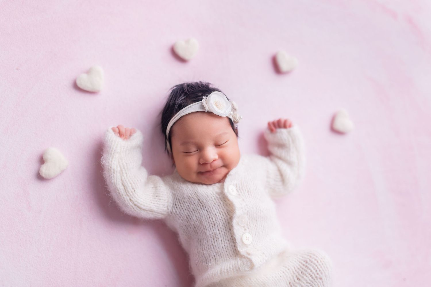 Baby in white outfit sleeping peacefully on pink backdrop with scattered decorative elements.