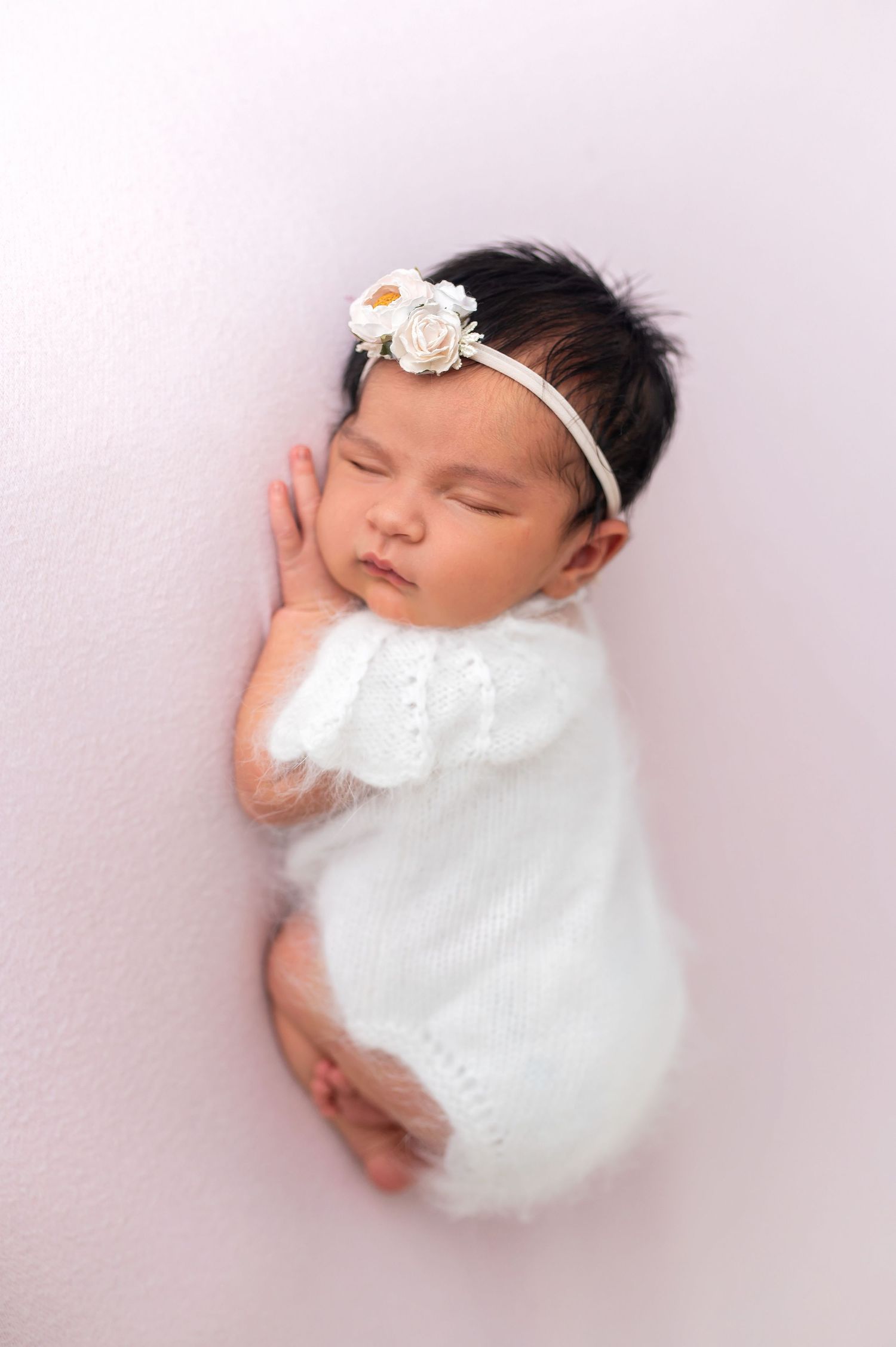Peaceful newborn wearing white outfit and floral headband sleeps on soft pink backdrop.