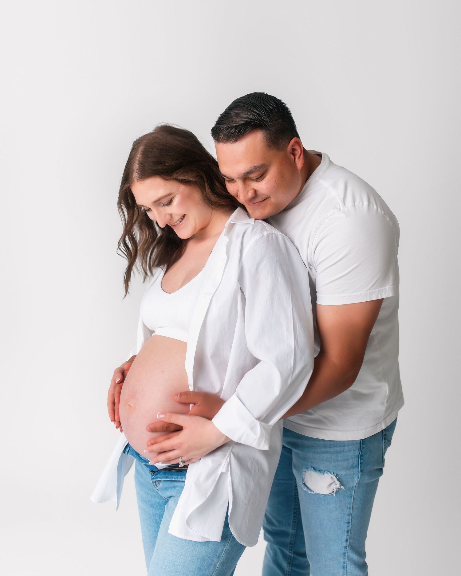 Couple in casual wear embrace while showing pregnant belly against white backdrop.