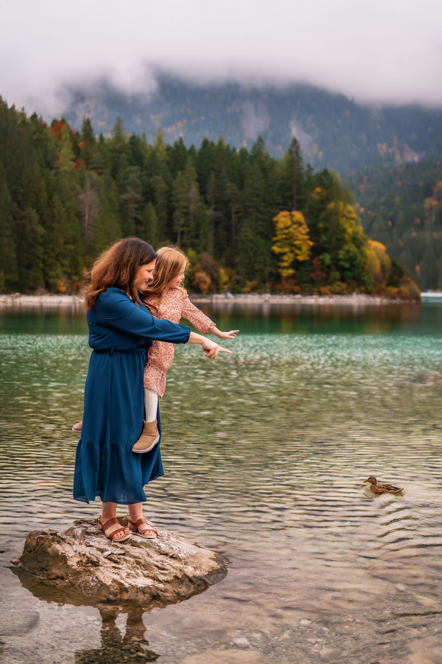 Two people in blue and pink dresses stand together on a rock in a scenic mountain lake with evergreen trees.