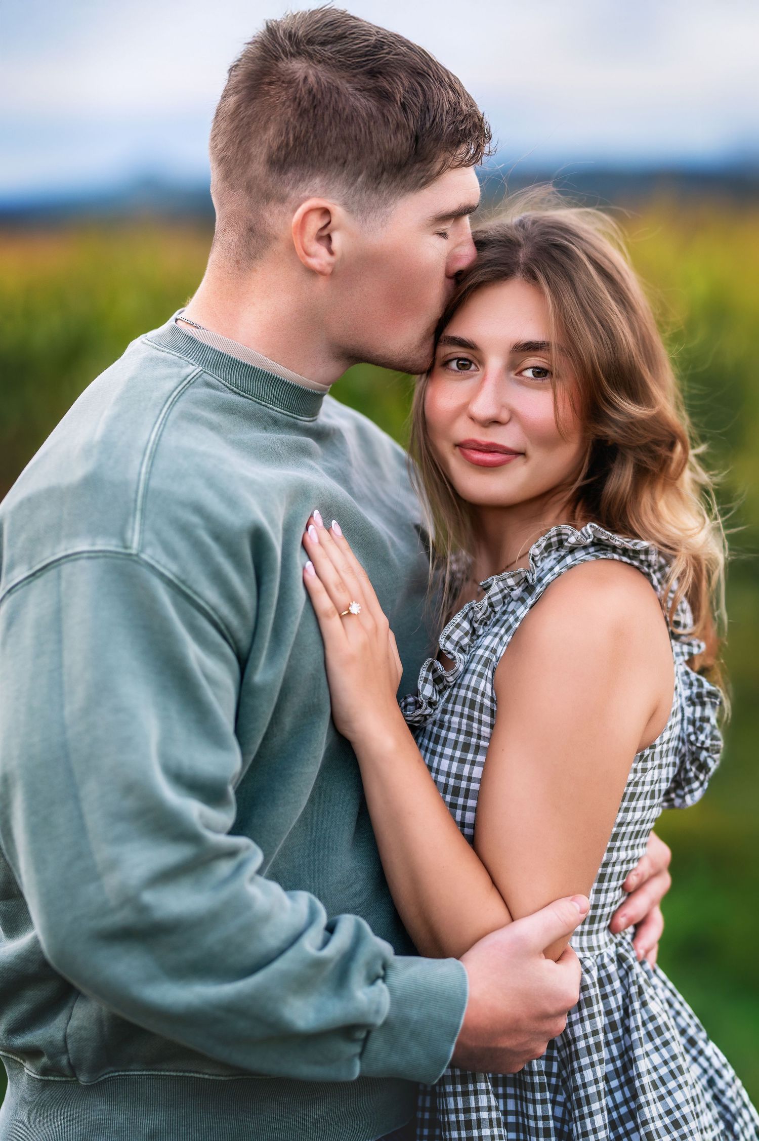 A couple embraces in a romantic outdoor photoshoot series against a scenic rural backdrop wearing green and gingham outfits.