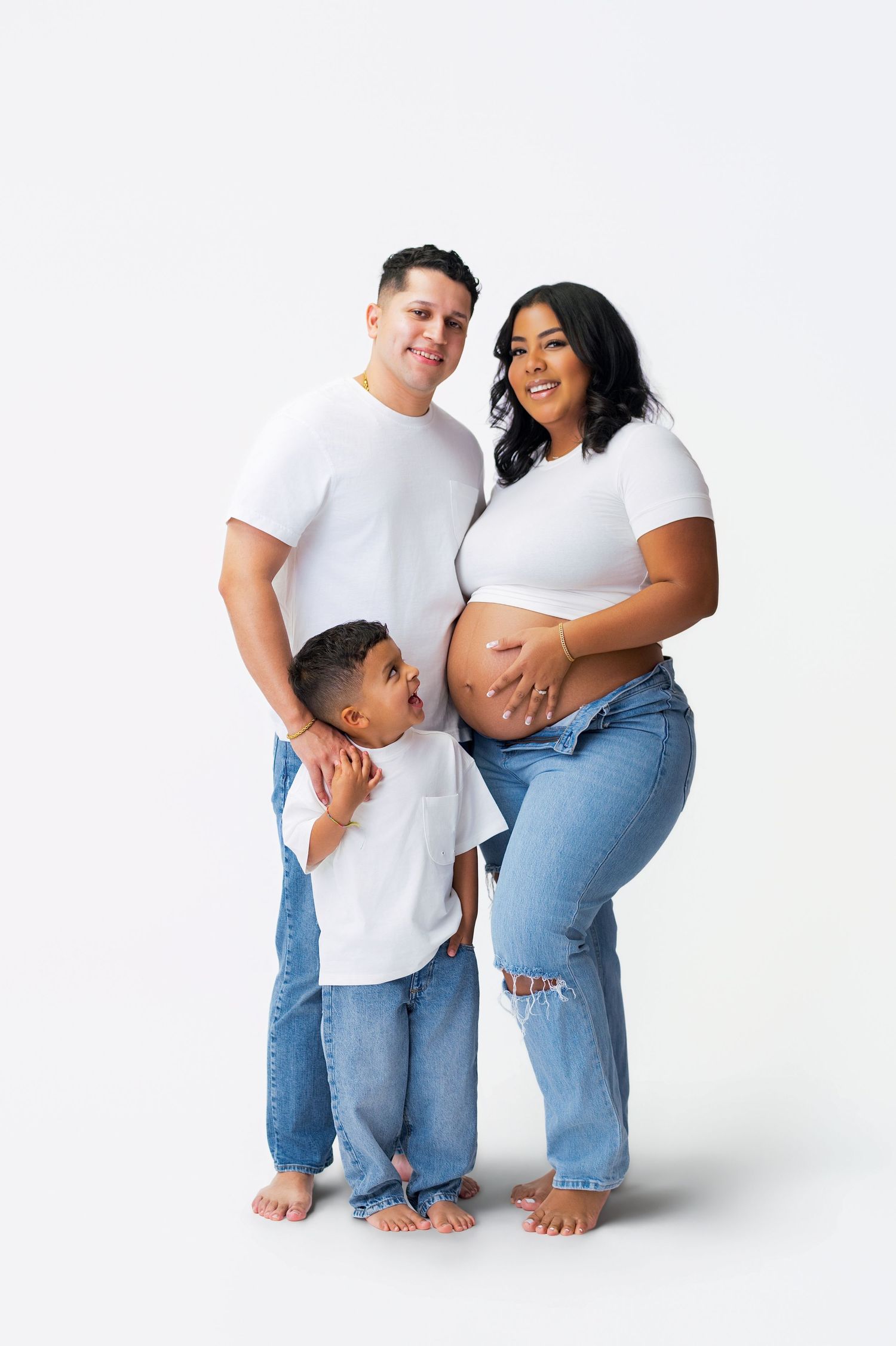 A family in matching white shirts and jeans poses together against a white backdrop for a maternity photo shoot.