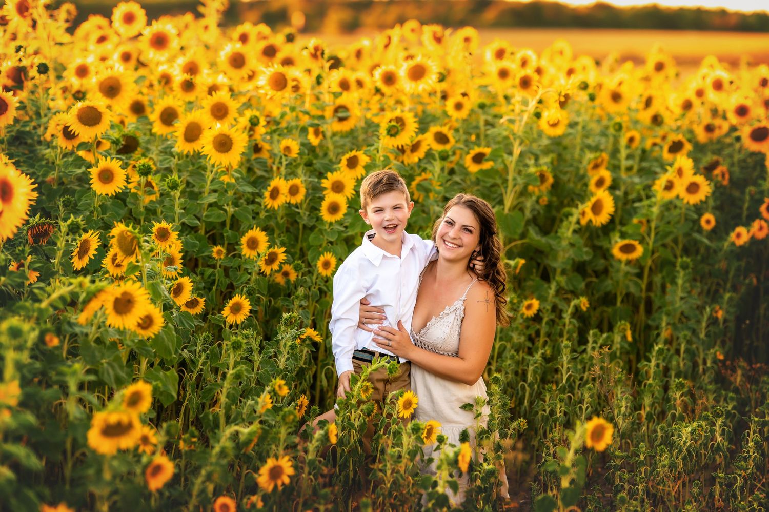 Woman and young boy smiling together in a vibrant sunflower field during golden hour sunset.