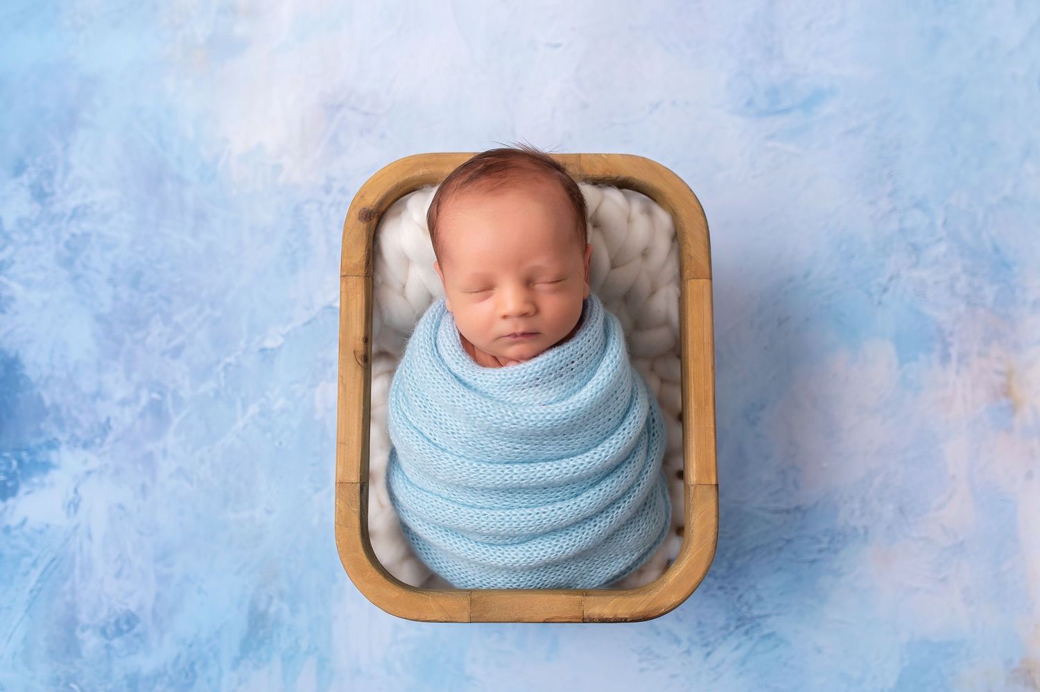 Sleeping newborn swaddled in blue wrap inside wooden basket on blue background.