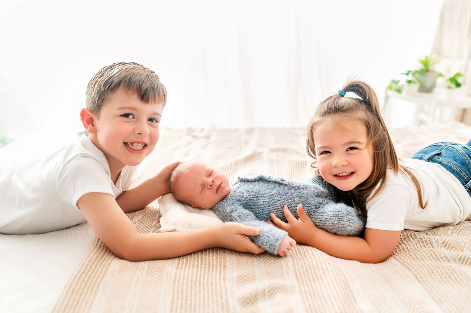 Two siblings lying on floor smiling while cuddling with newborn baby on beige carpet.