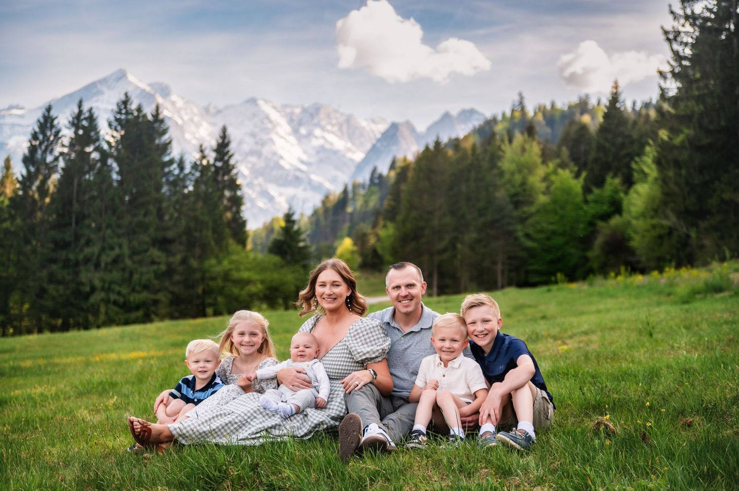 Family of six posing on green grass with snow-capped mountains and pine trees in the background.